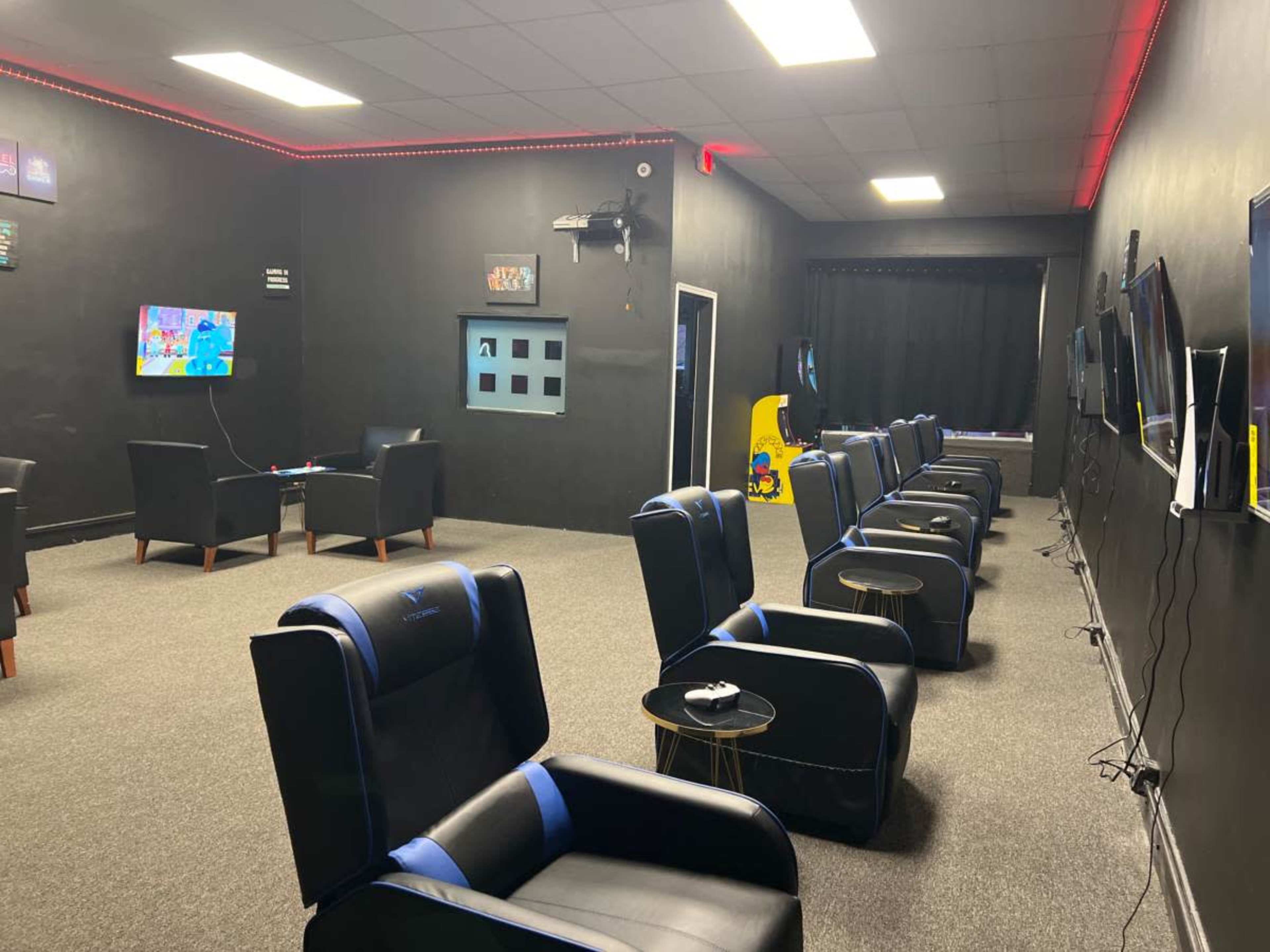 A gaming lounge with several black and blue chairs arranged in rows facing wall-mounted televisions, along with arcade games at one end of the room.
