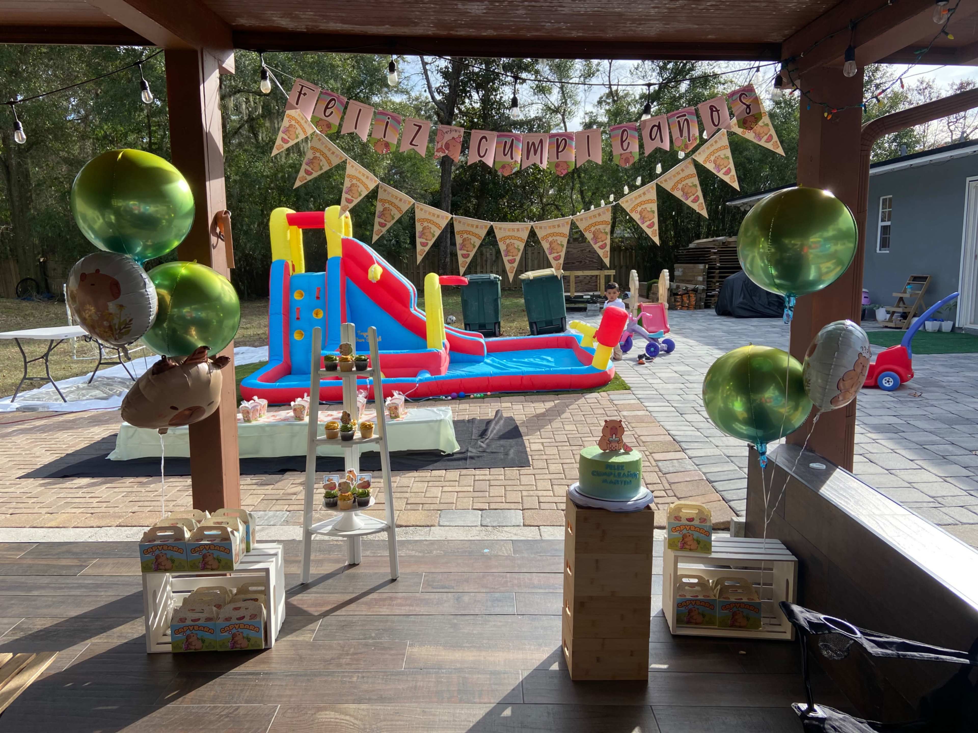 The image shows a children's birthday party setup with a colorful inflatable slide in the background, a "Feliz cumpleaños" banner, and various decorations including balloons and a cake.