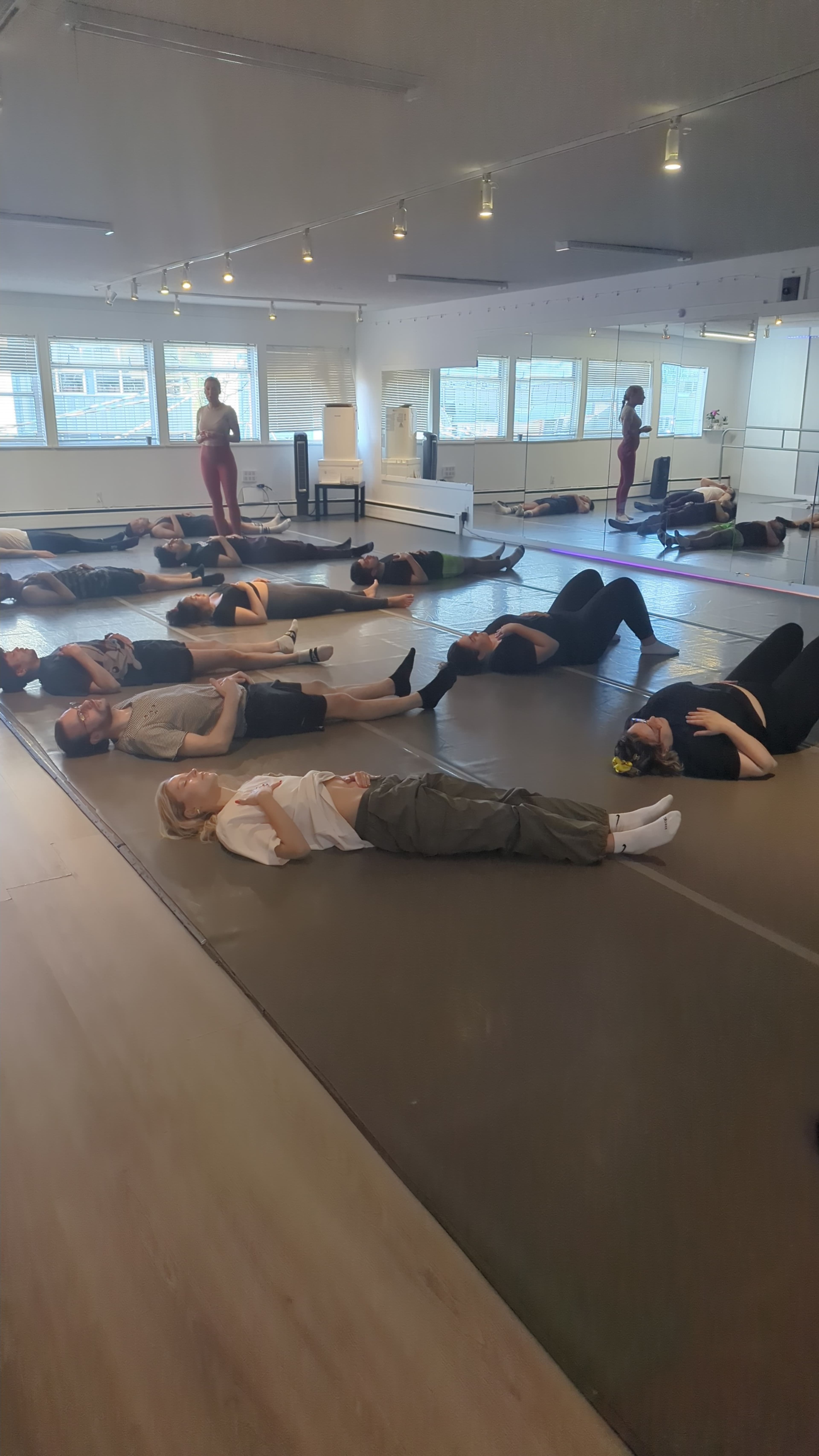 A group of participants is lying on mats in a spacious studio, engaged in a floor exercise while others stand nearby.