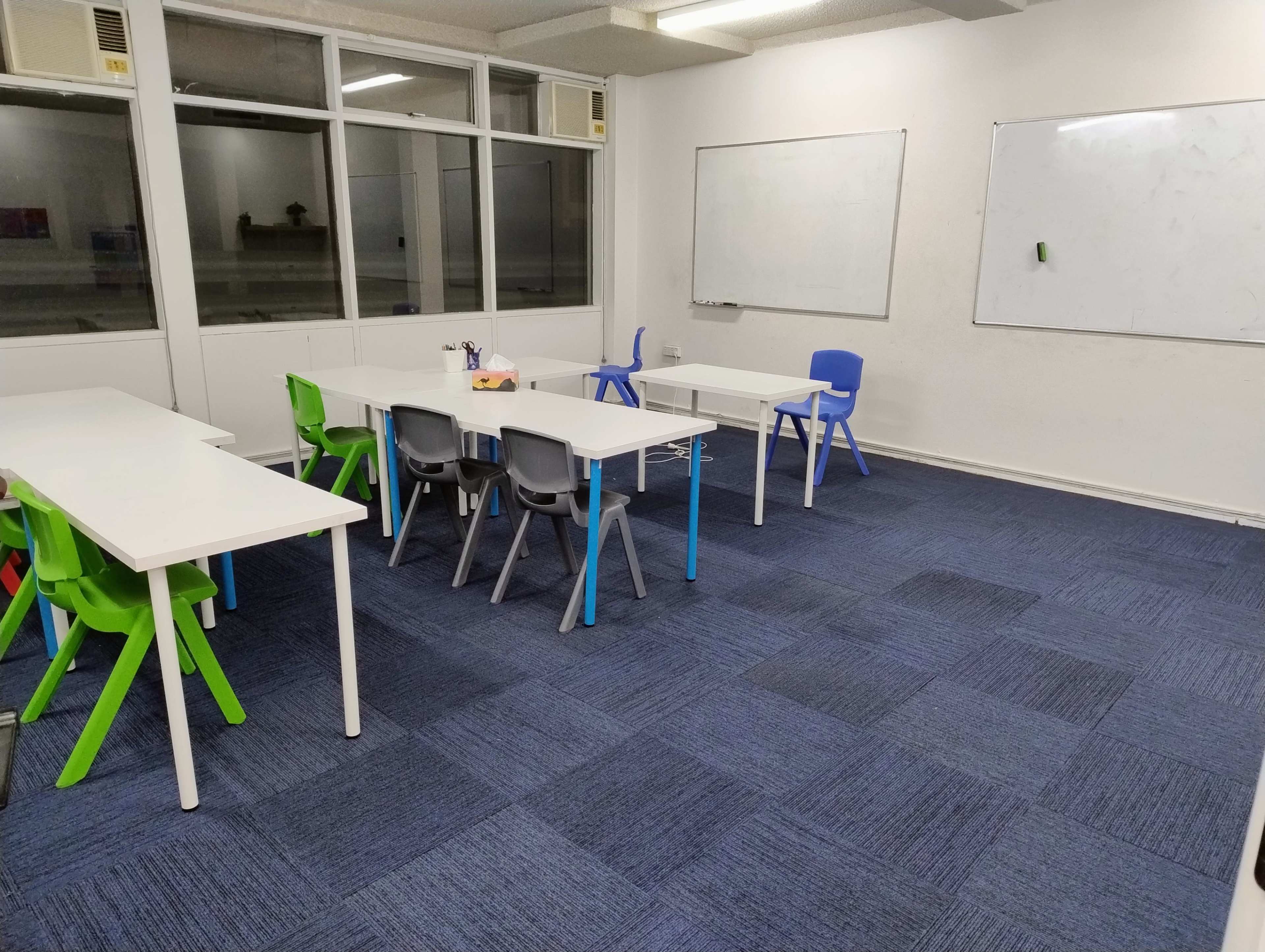 A classroom with white tables and colorful chairs arranged in a simple layout, featuring windows and whiteboards on the walls.