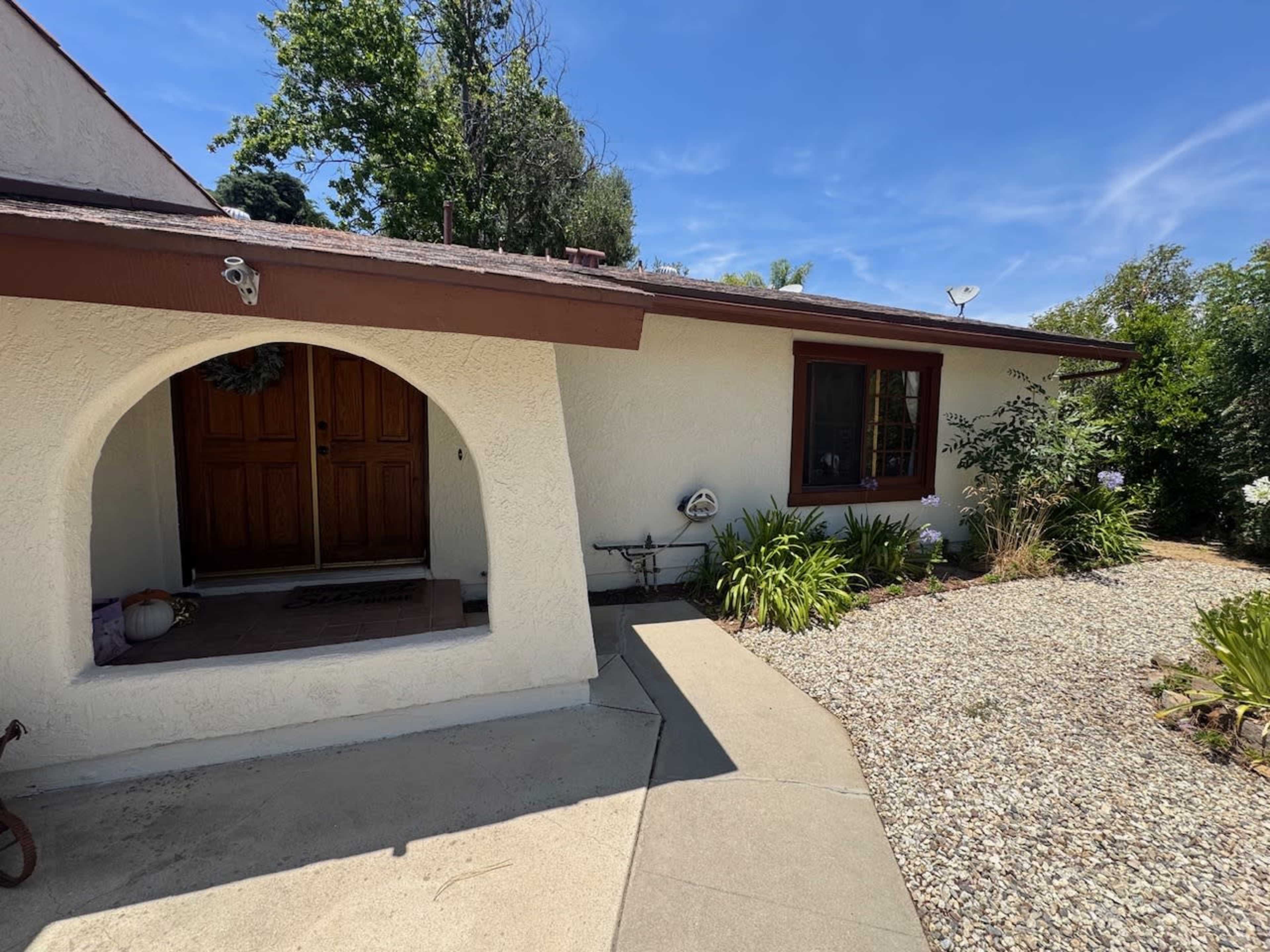 The image shows the entrance of a house featuring a covered porch, two wooden doors, and a landscaped path lined with gravel and plants.