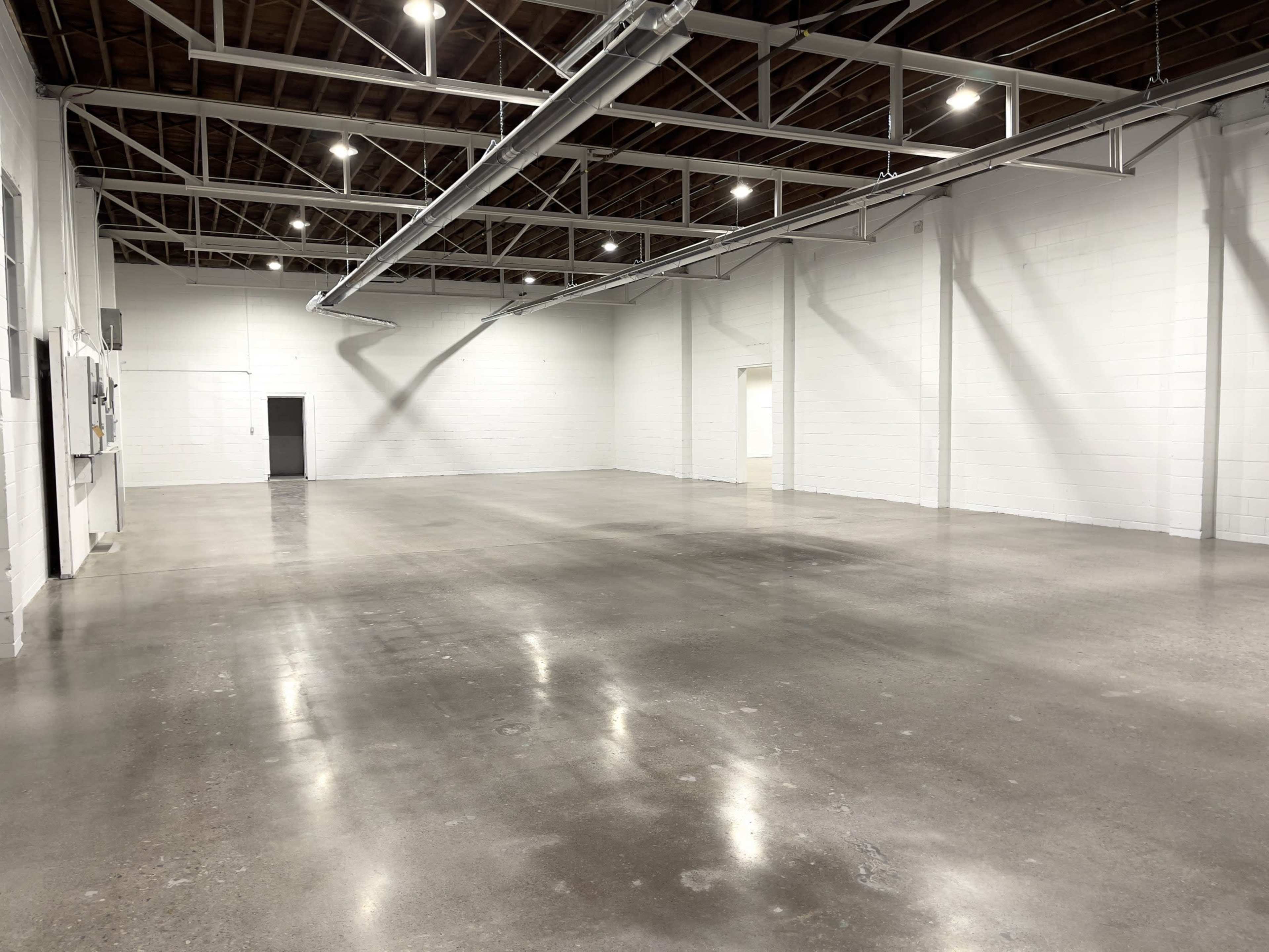 The image shows a spacious, empty room with polished concrete flooring and white walls, illuminated by overhead lights.