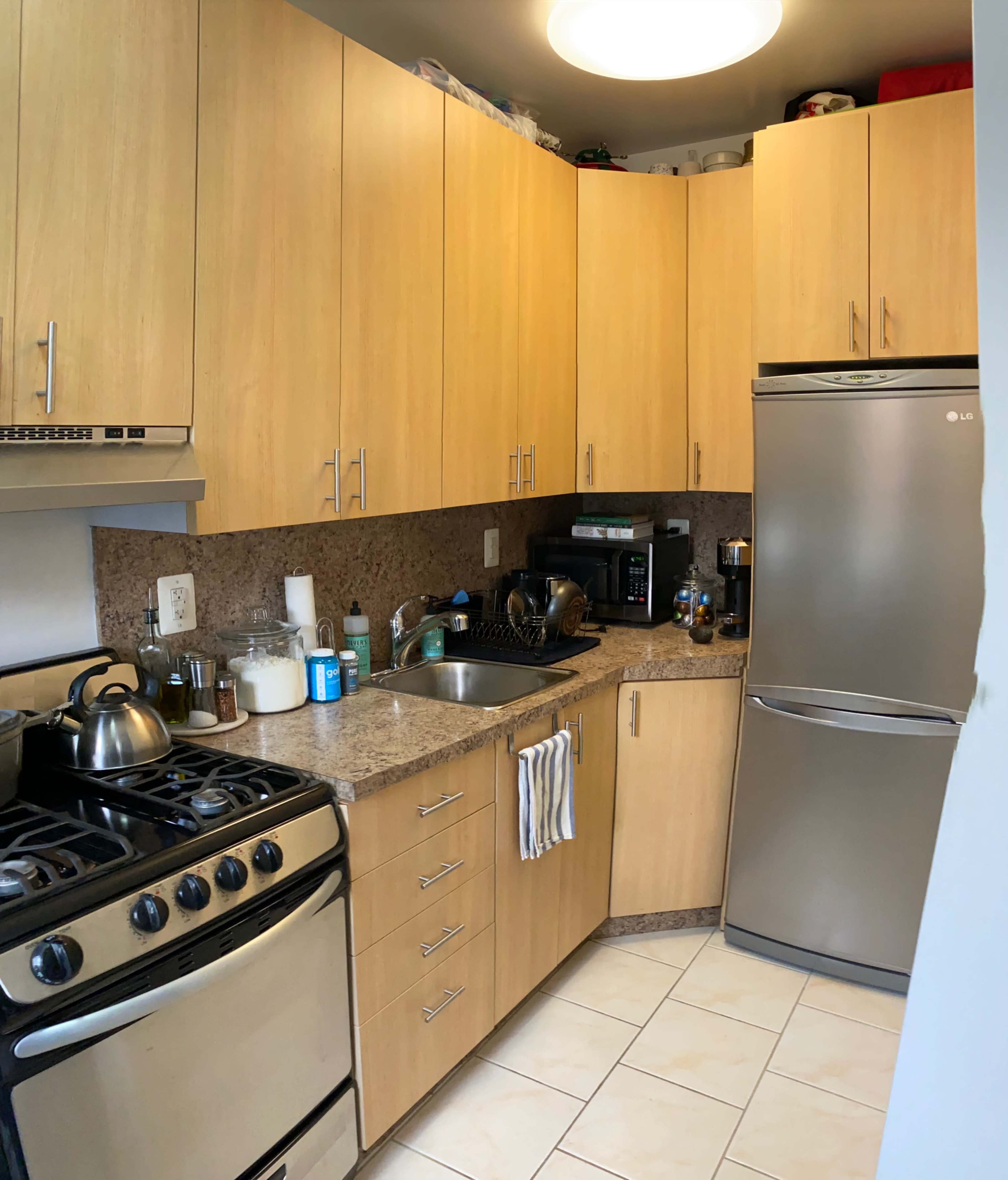 A compact kitchen with wooden cabinets, a stainless steel refrigerator, a gas stove, and a sink, all arranged along a tiled floor.