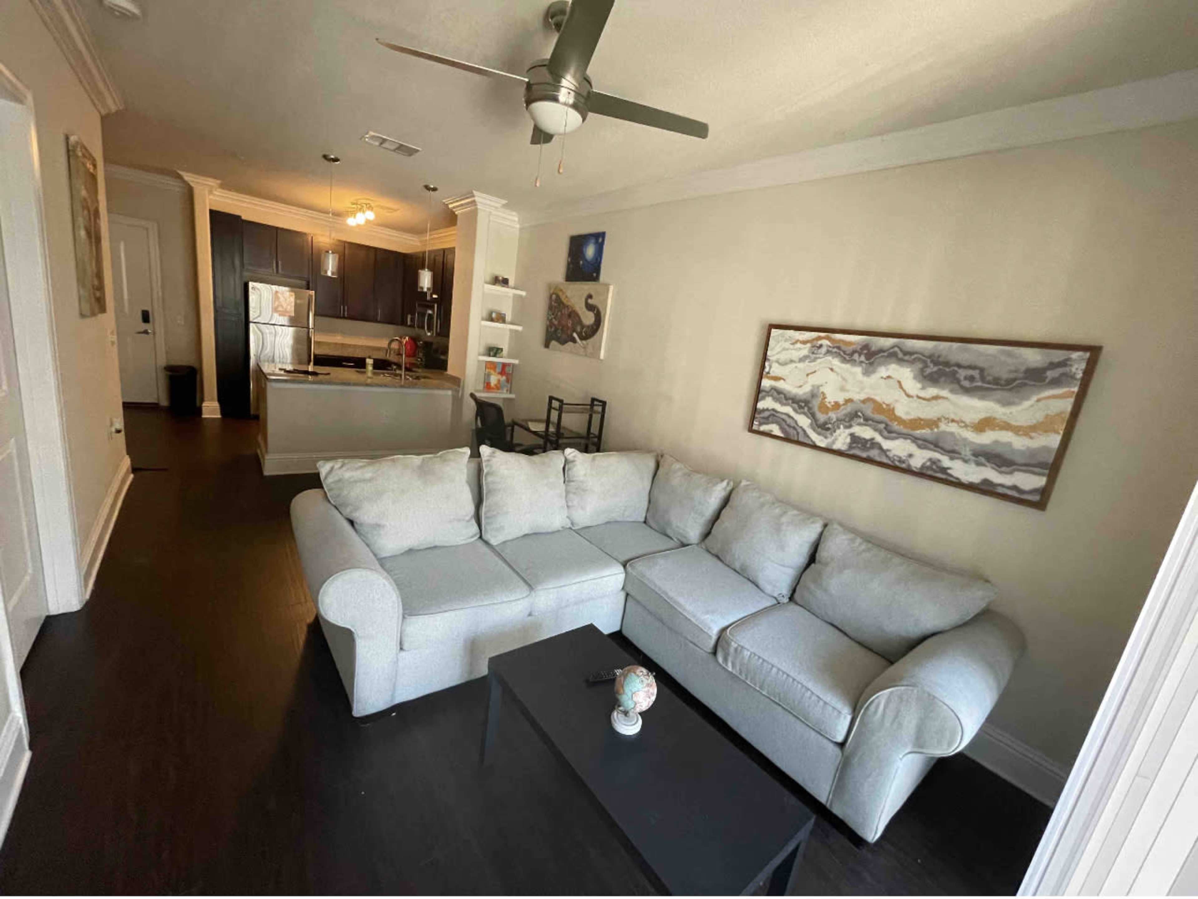 The image shows a living room with a light gray sectional sofa, a black coffee table, and an open kitchen in the background.
