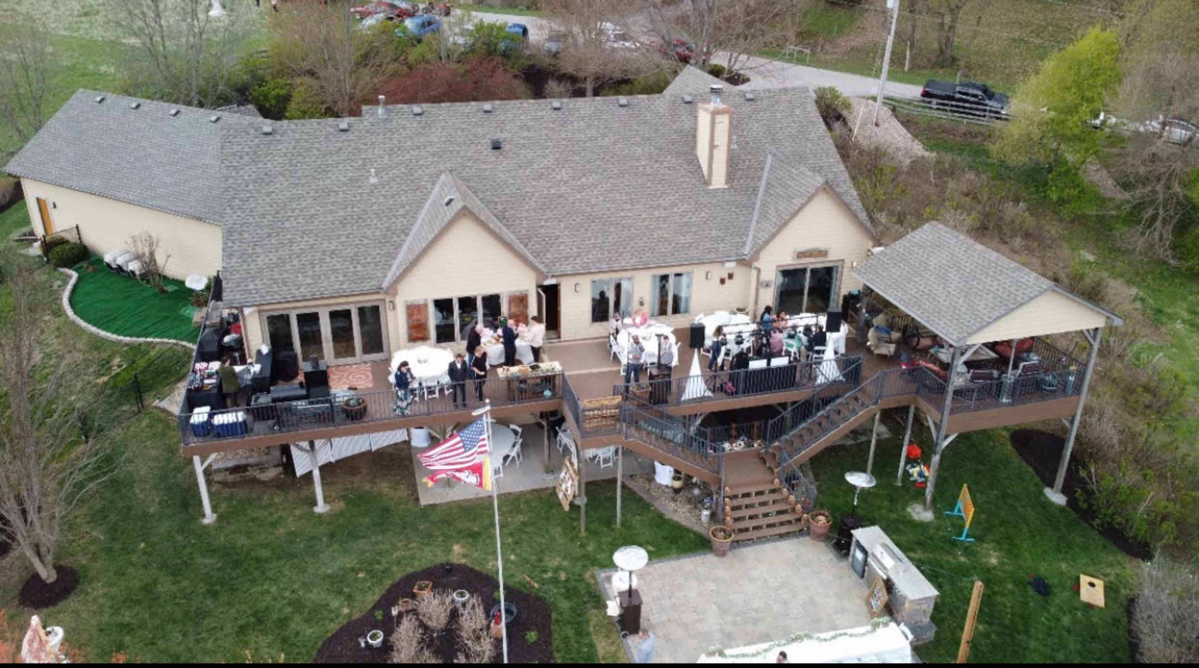 The image shows a spacious two-story house with a large deck, where a gathering of people is taking place, along with a well-manicured lawn and various outdoor decorations.