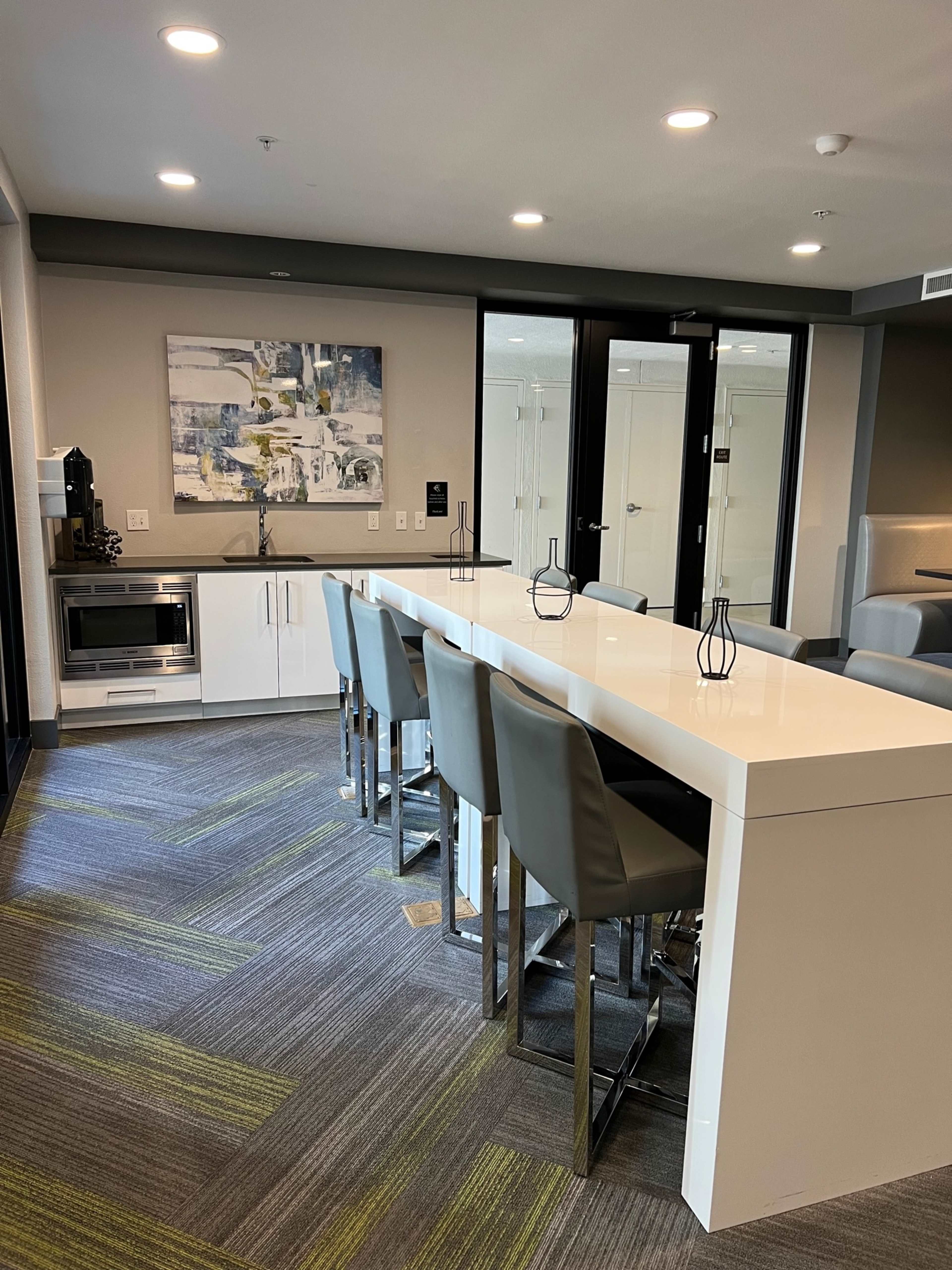 The image shows a modern kitchen area with a long white table surrounded by gray high-backed chairs, a microwave, and a small kitchenette on the side.