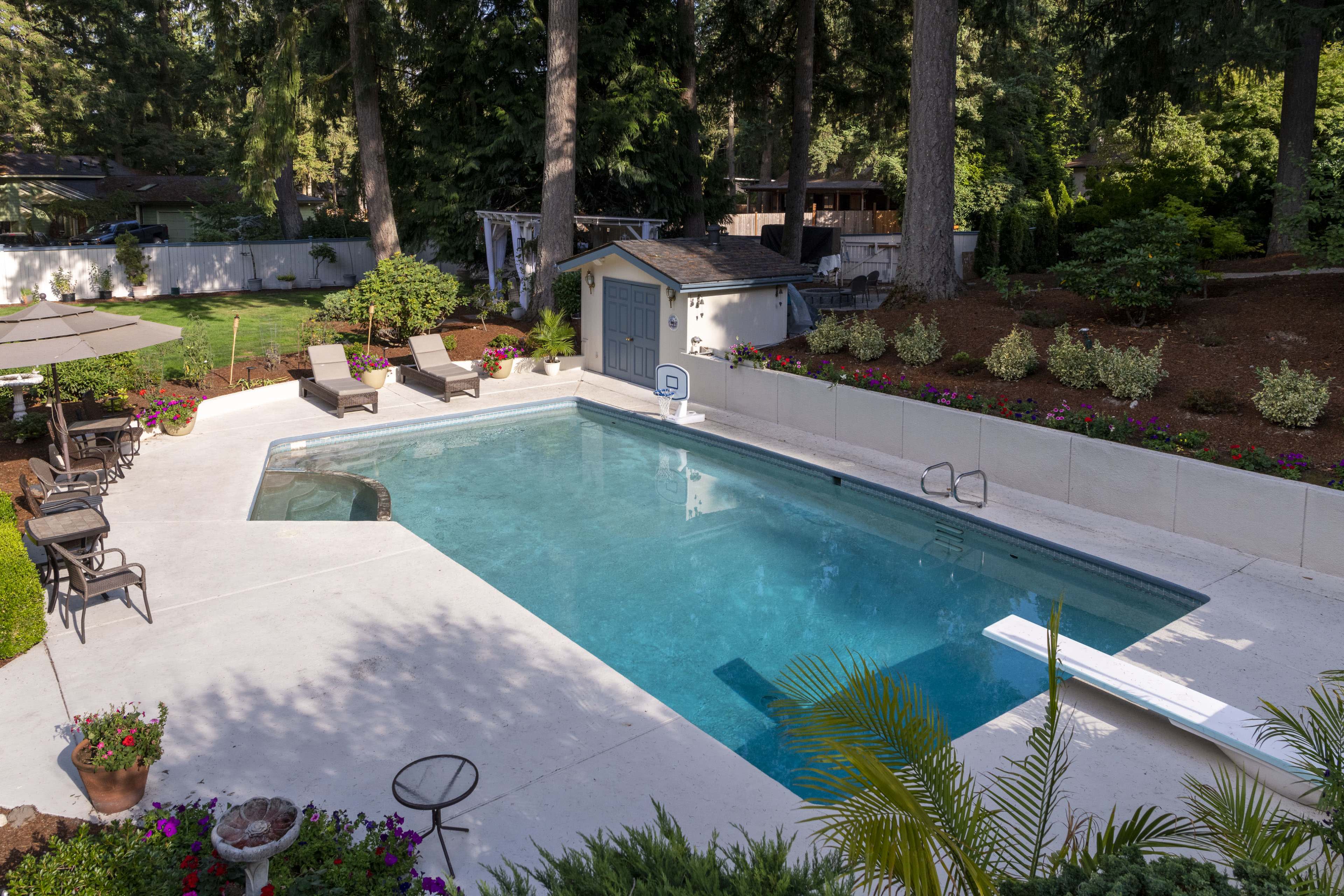 A rectangular swimming pool surrounded by lounge chairs, a diving board, and landscaped gardens with trees in the background.