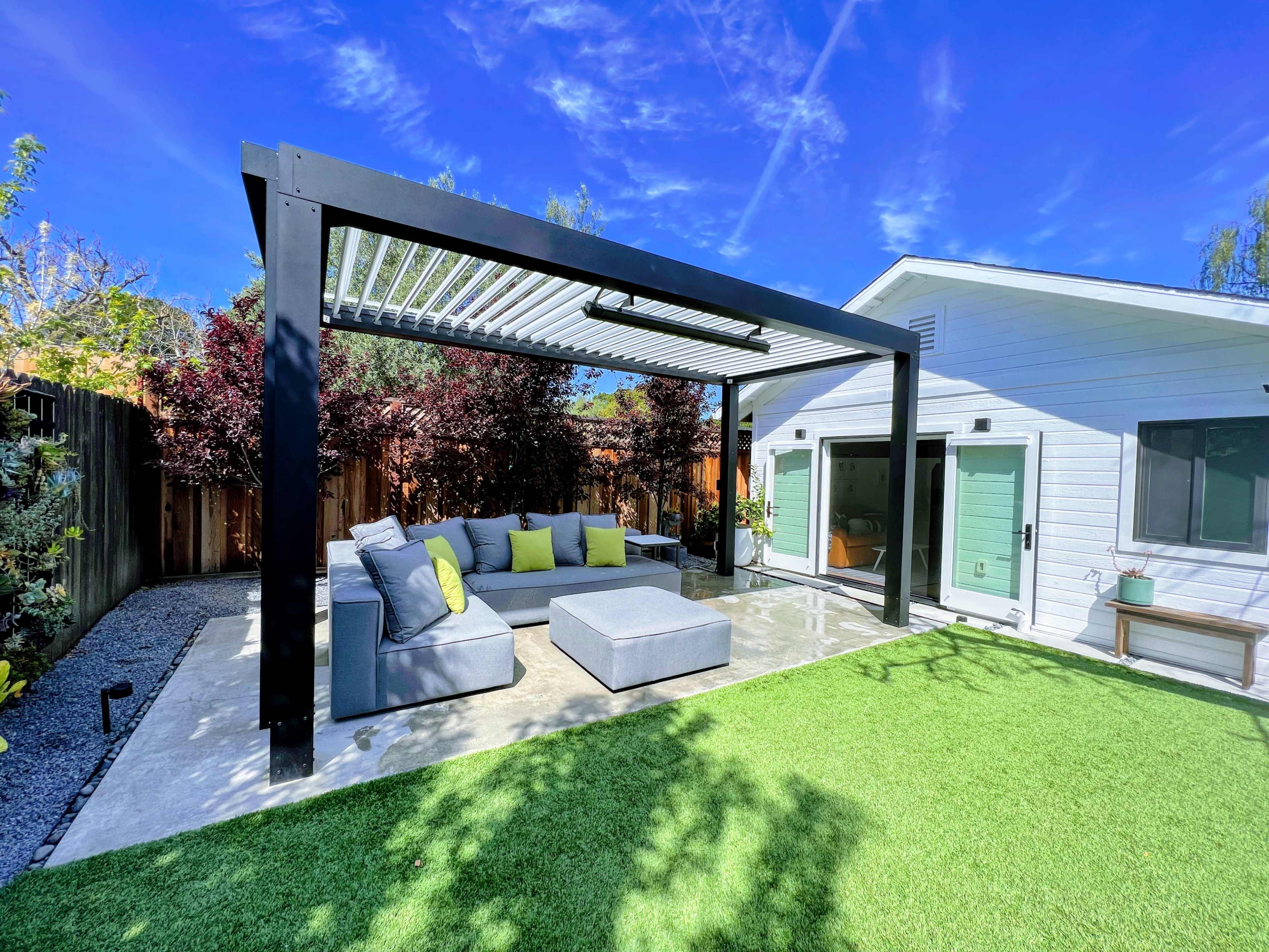 A modern outdoor lounging area with a pergola, featuring a sectional sofa and coffee table on a grassy lawn, adjacent to a white house.