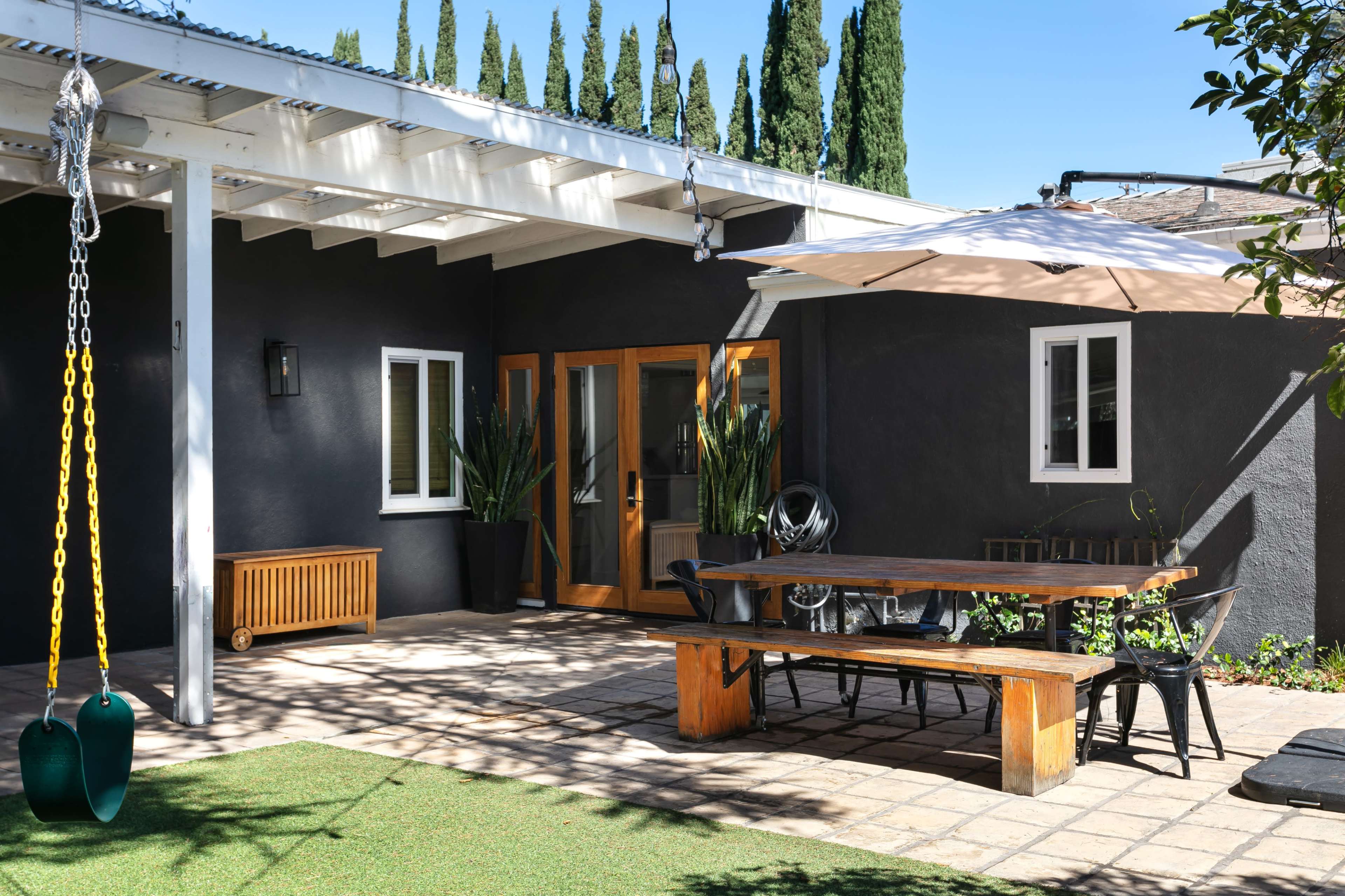 A patio features a large wooden dining table, an umbrella, and a swing set, surrounded by greenery and modern architecture.