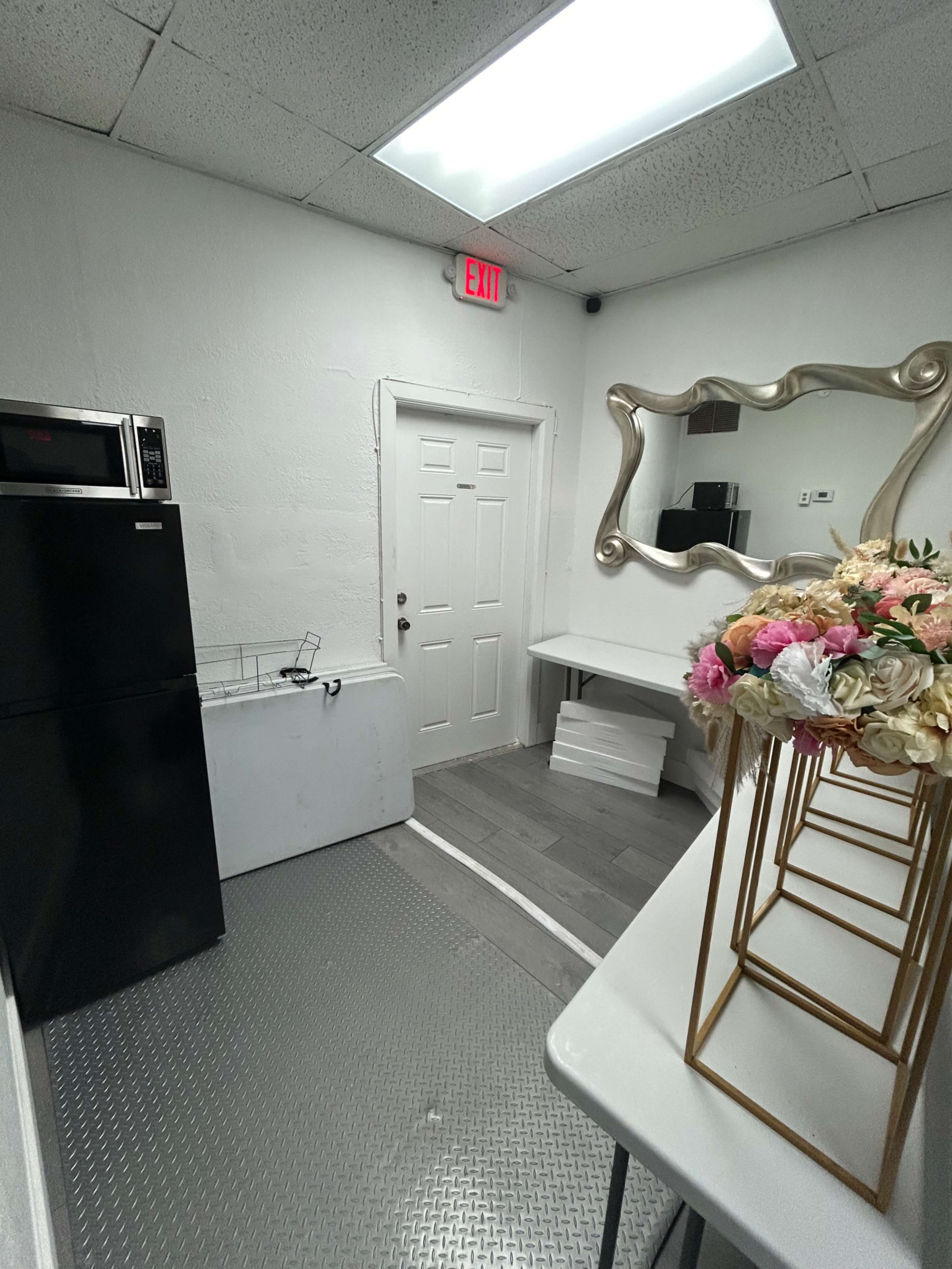 The image shows a small room with a refrigerator, a freezer, a table, and a mirror on the wall, featuring a floral arrangement on a stand.