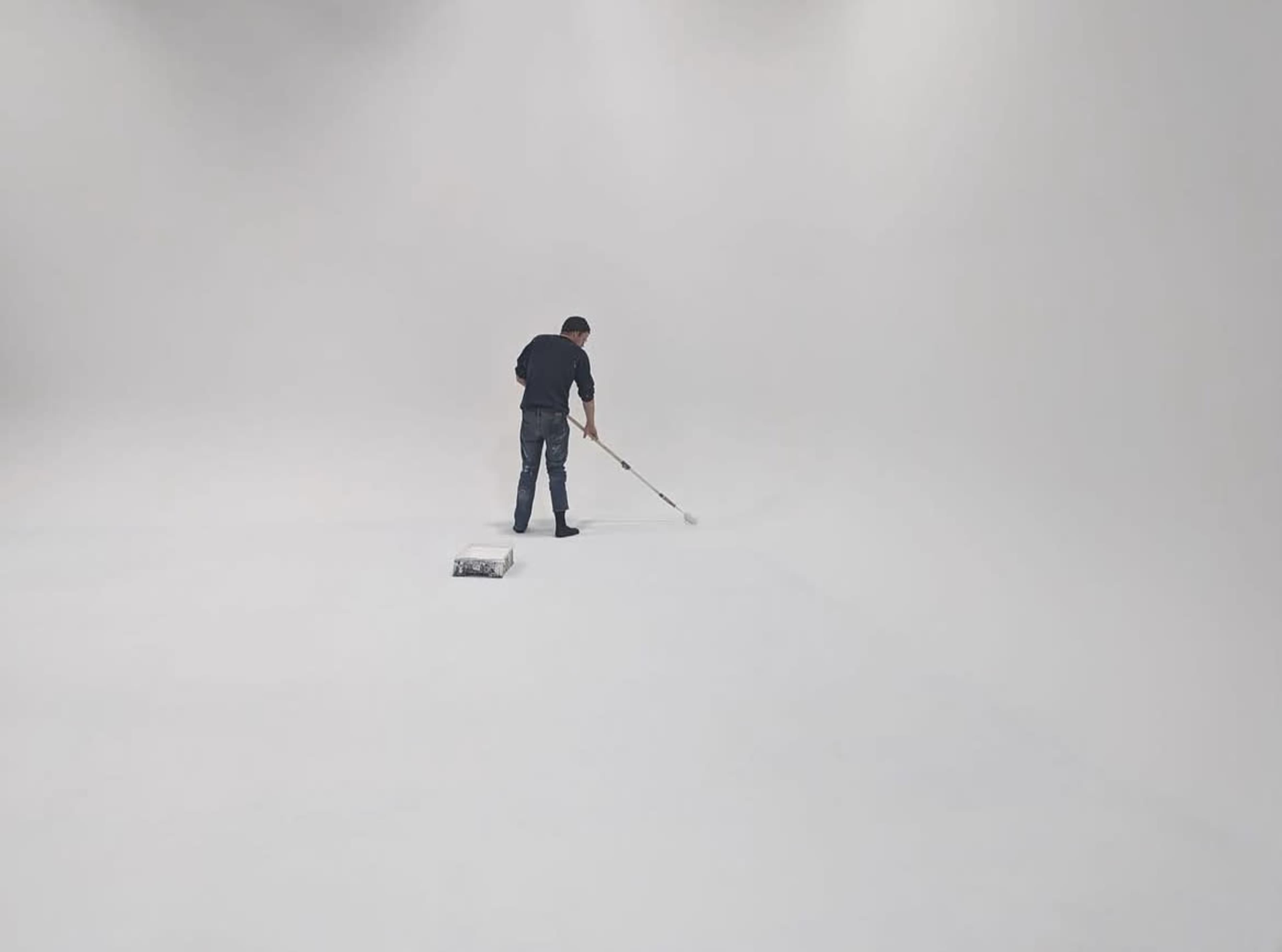 A person uses a paint roller to apply white paint on a large, empty studio floor.