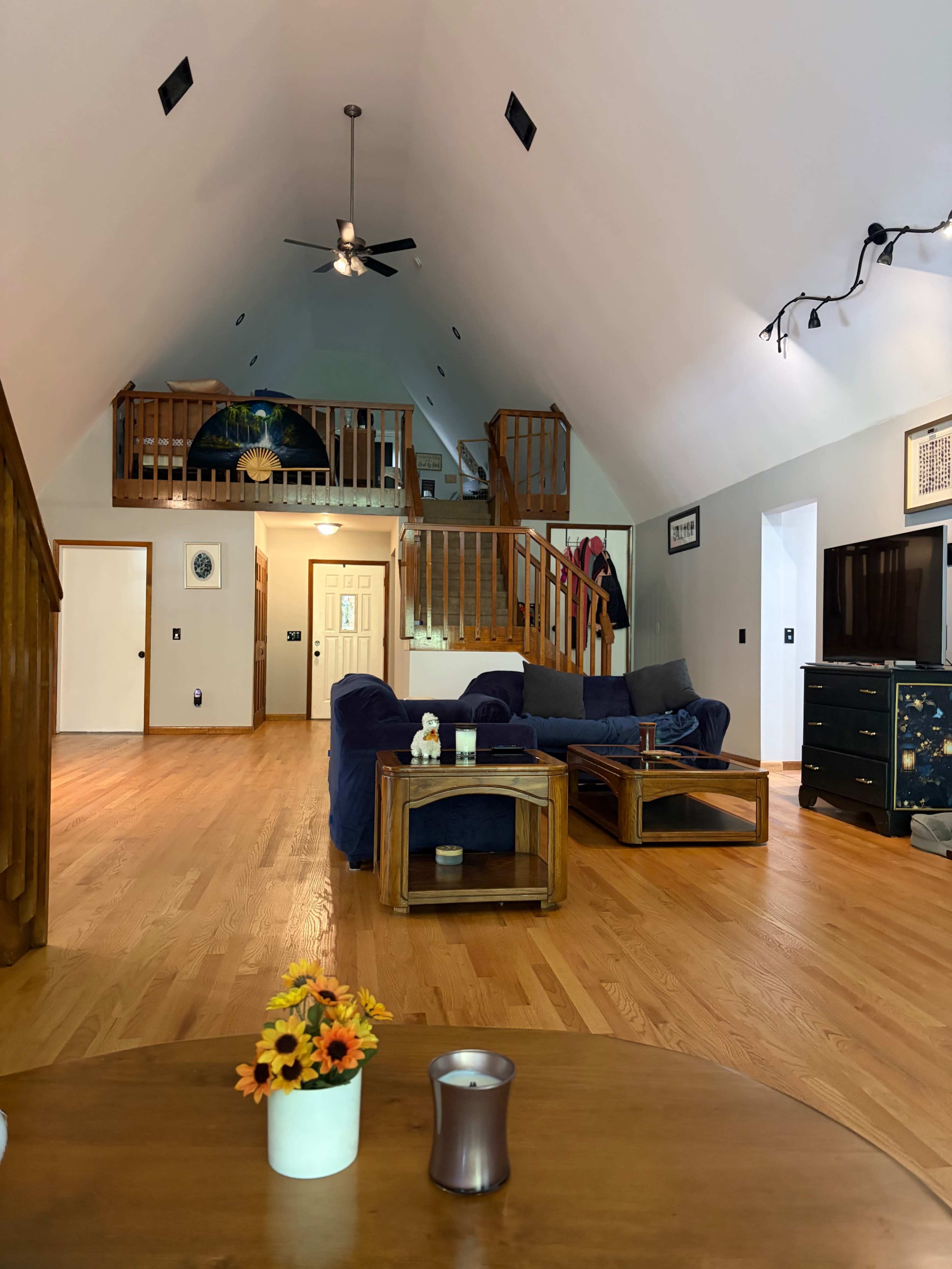 The image shows a spacious living area with a high ceiling, wooden floors, and a sofa set, featuring a staircase leading to an upper level.
