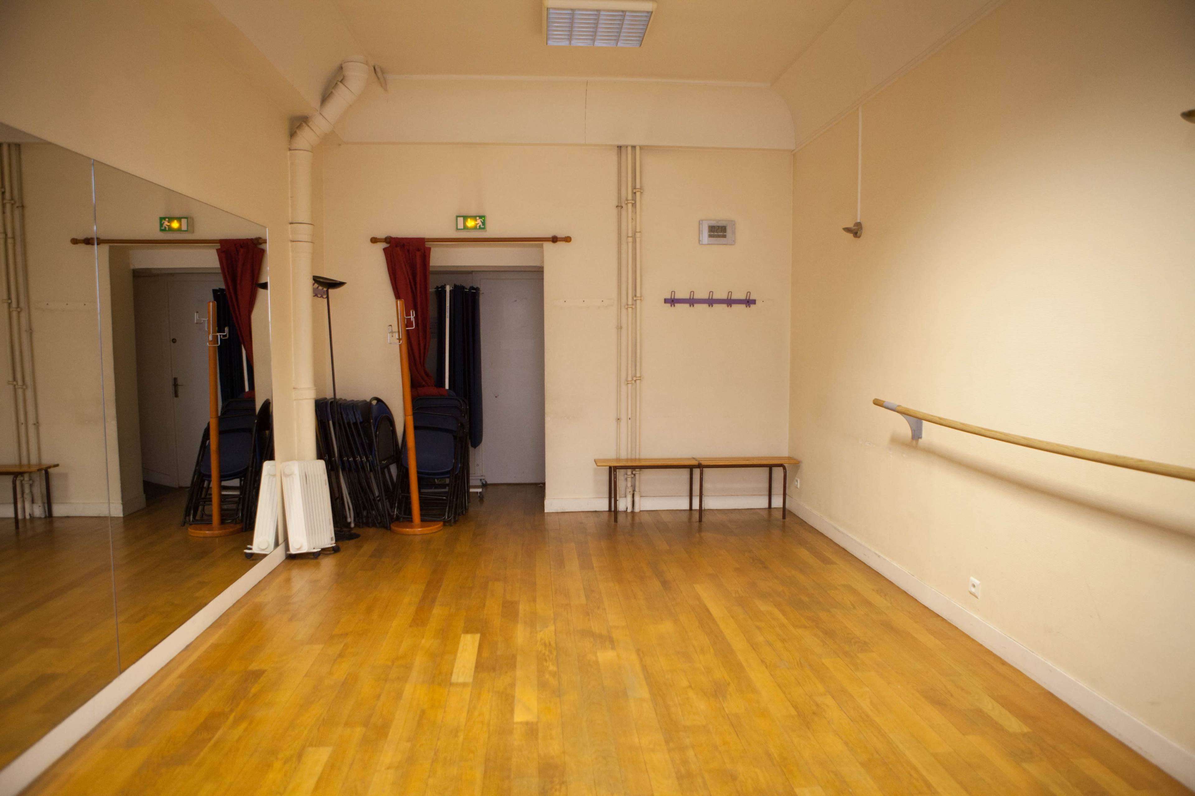 The image shows a wooden-floored dance studio with mirrors on one wall, a barre, a bench, and stacked chairs in the corner.