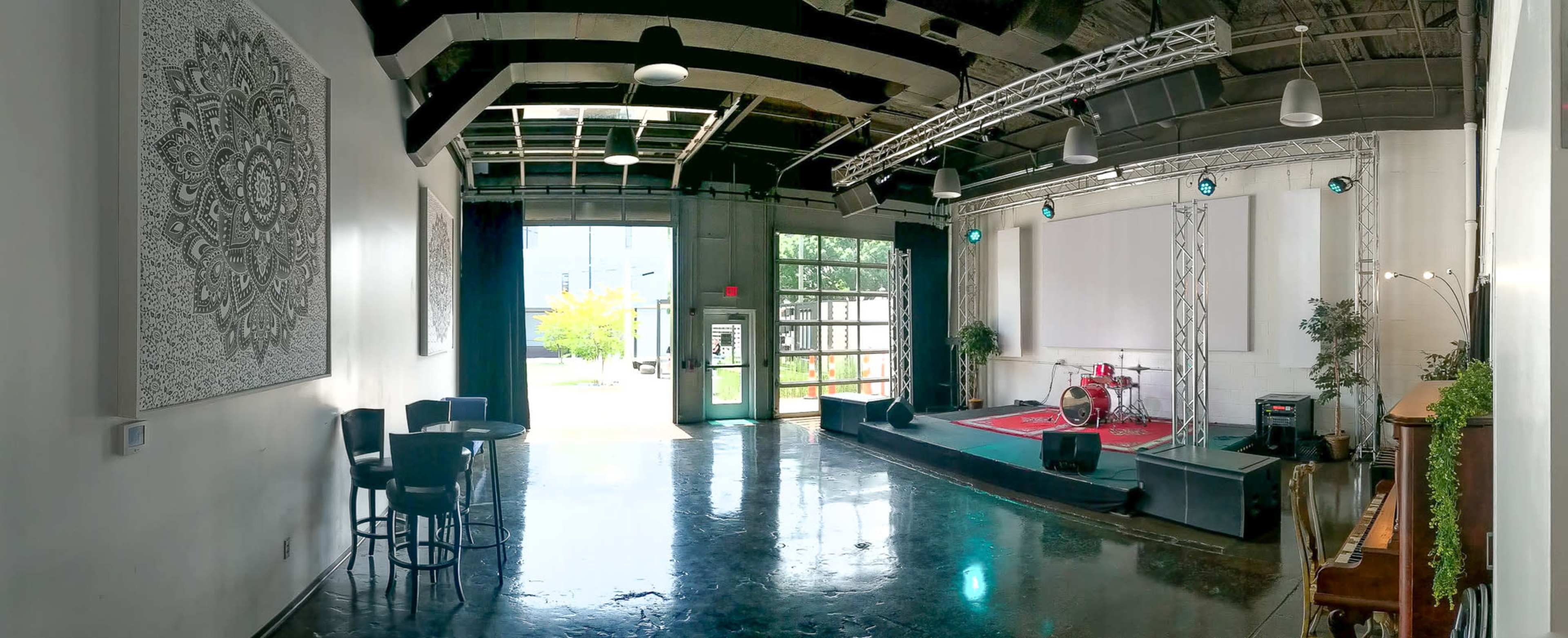The image shows a spacious indoor performance venue with a stage, drum set, and seating areas, featuring large windows that open to an outdoor area.