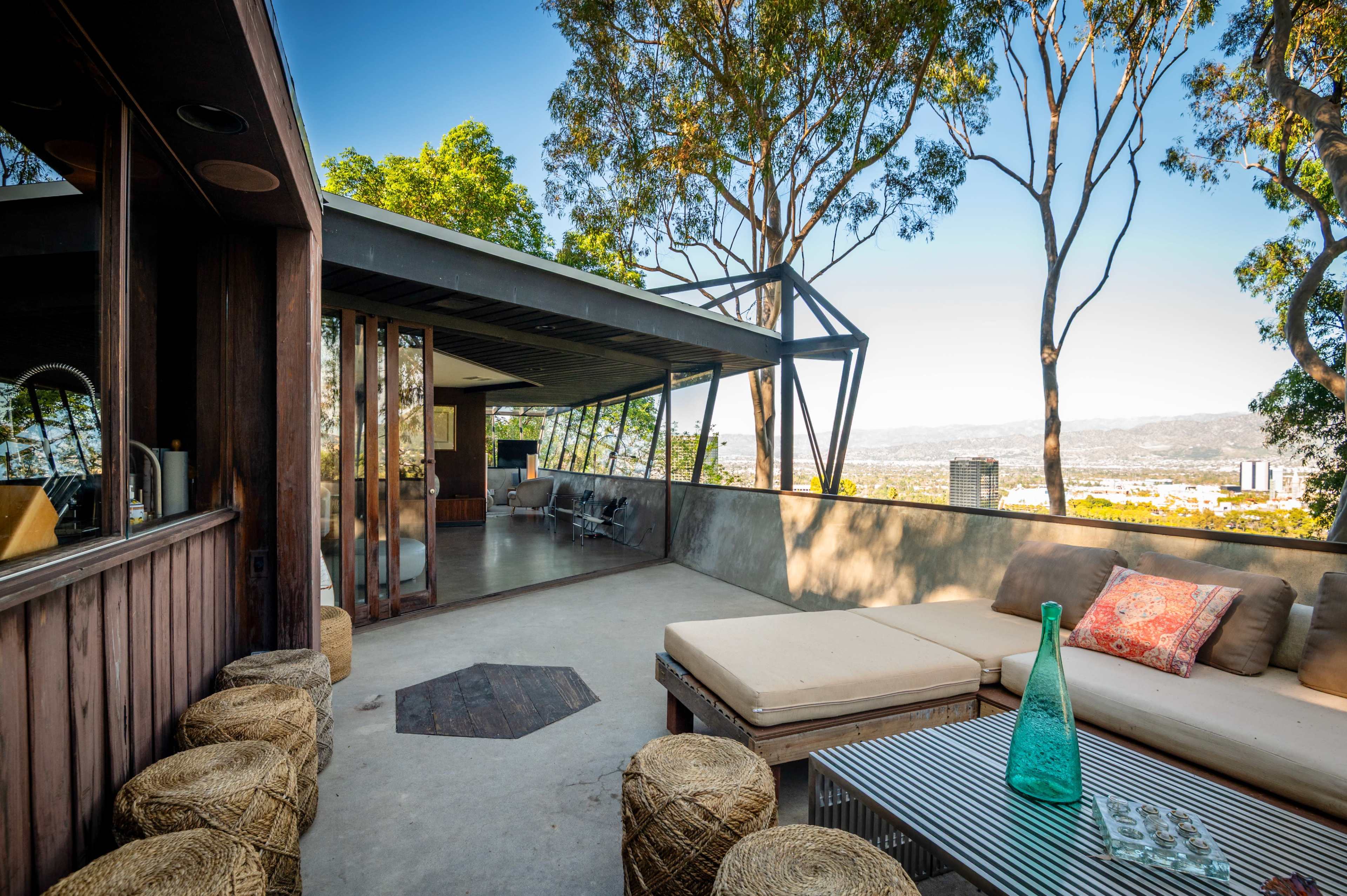 The image shows a modern outdoor lounge area with a sofa, a glass coffee table, and a view of a city nestled among green trees.