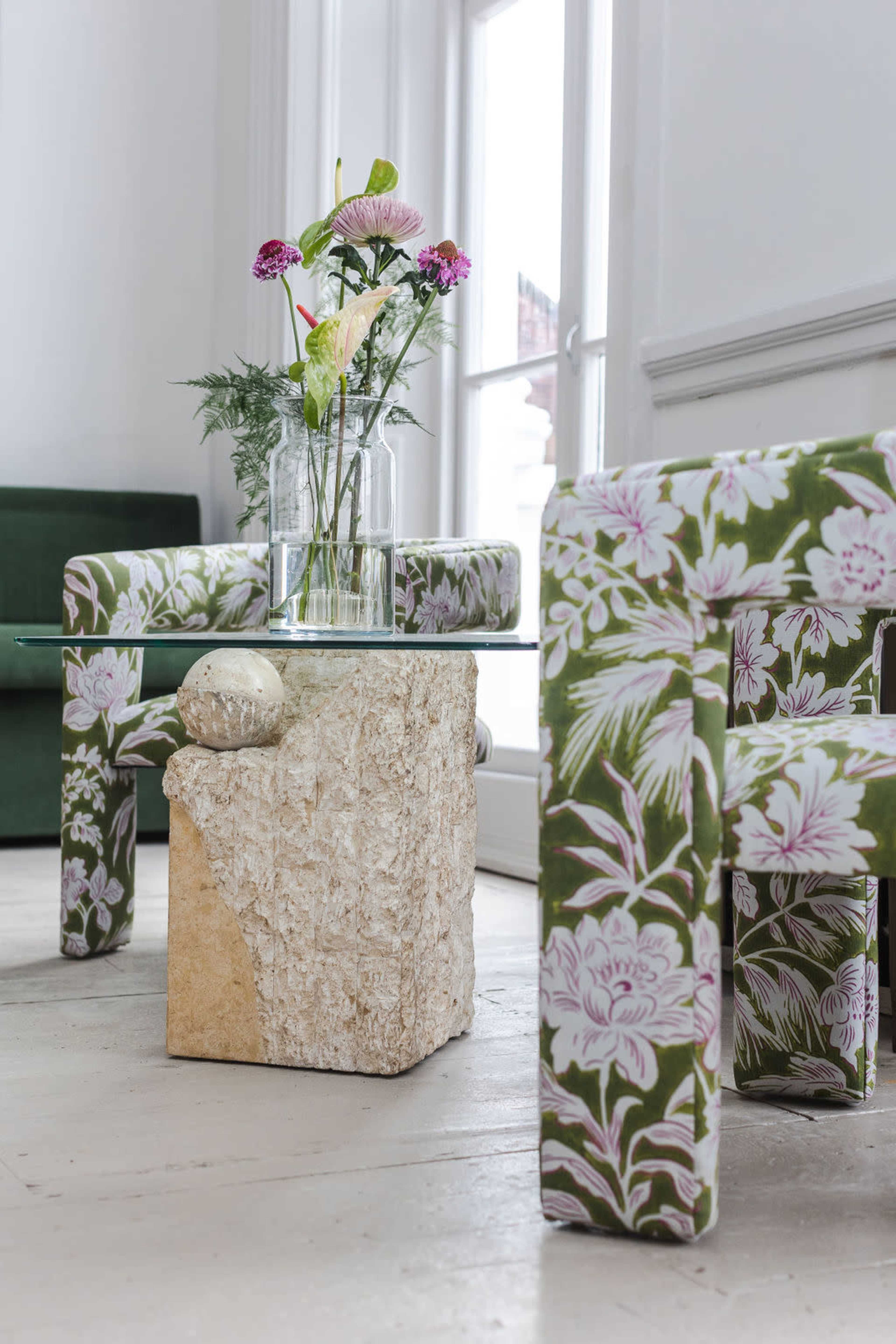 A pair of upholstered chairs with floral patterns facing a glass-topped stone coffee table adorned with a vase of flowers.