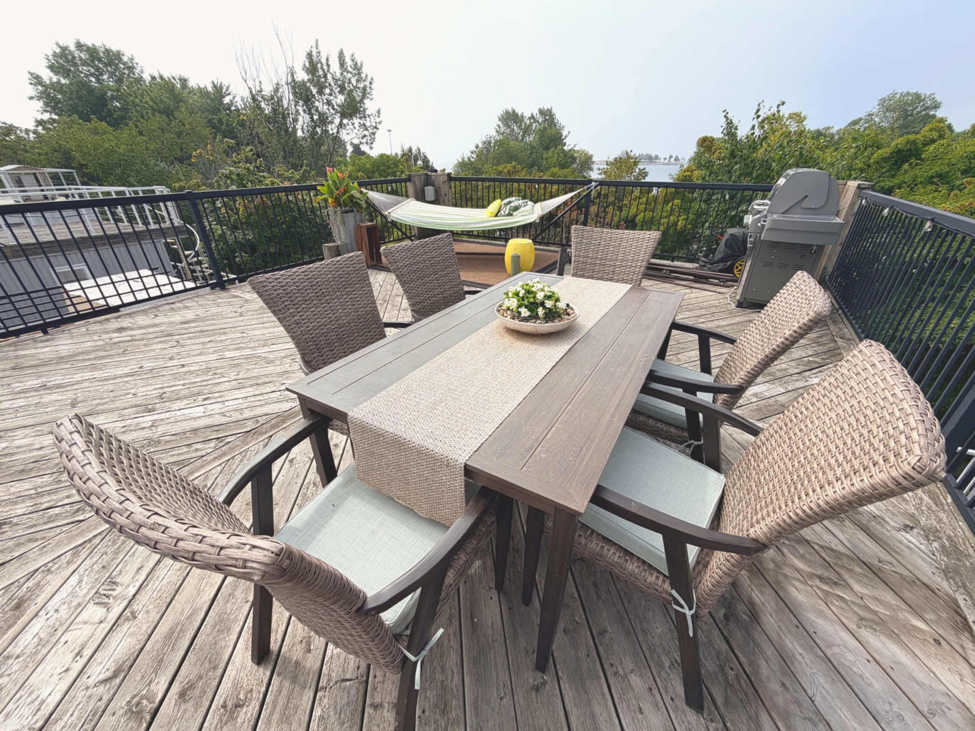 A wooden deck features a rectangular table surrounded by six woven chairs, with a centerpiece of flowers and a hammock visible in the background.