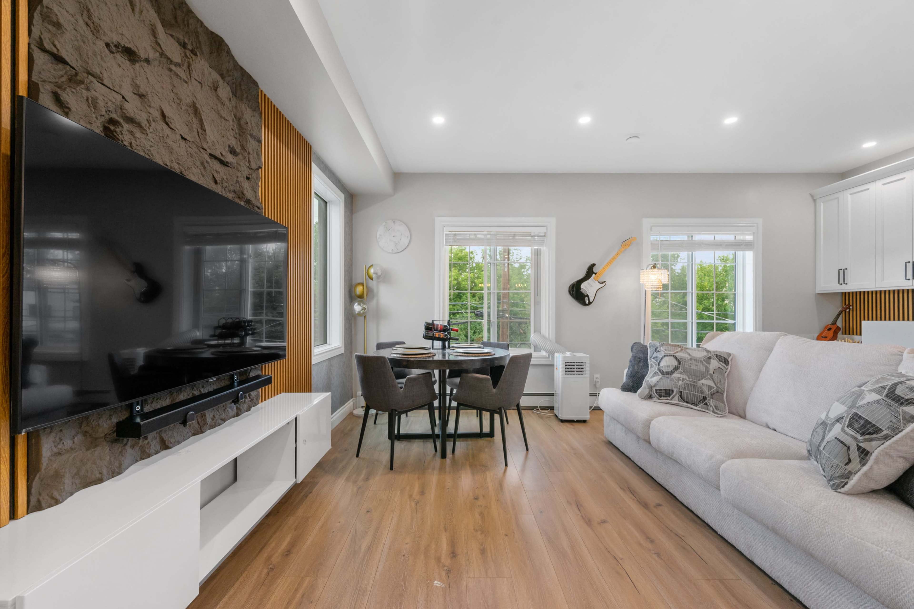 The scene features a modern living room with a large television mounted on a stone wall, a circular dining table, and a light-colored sectional sofa.