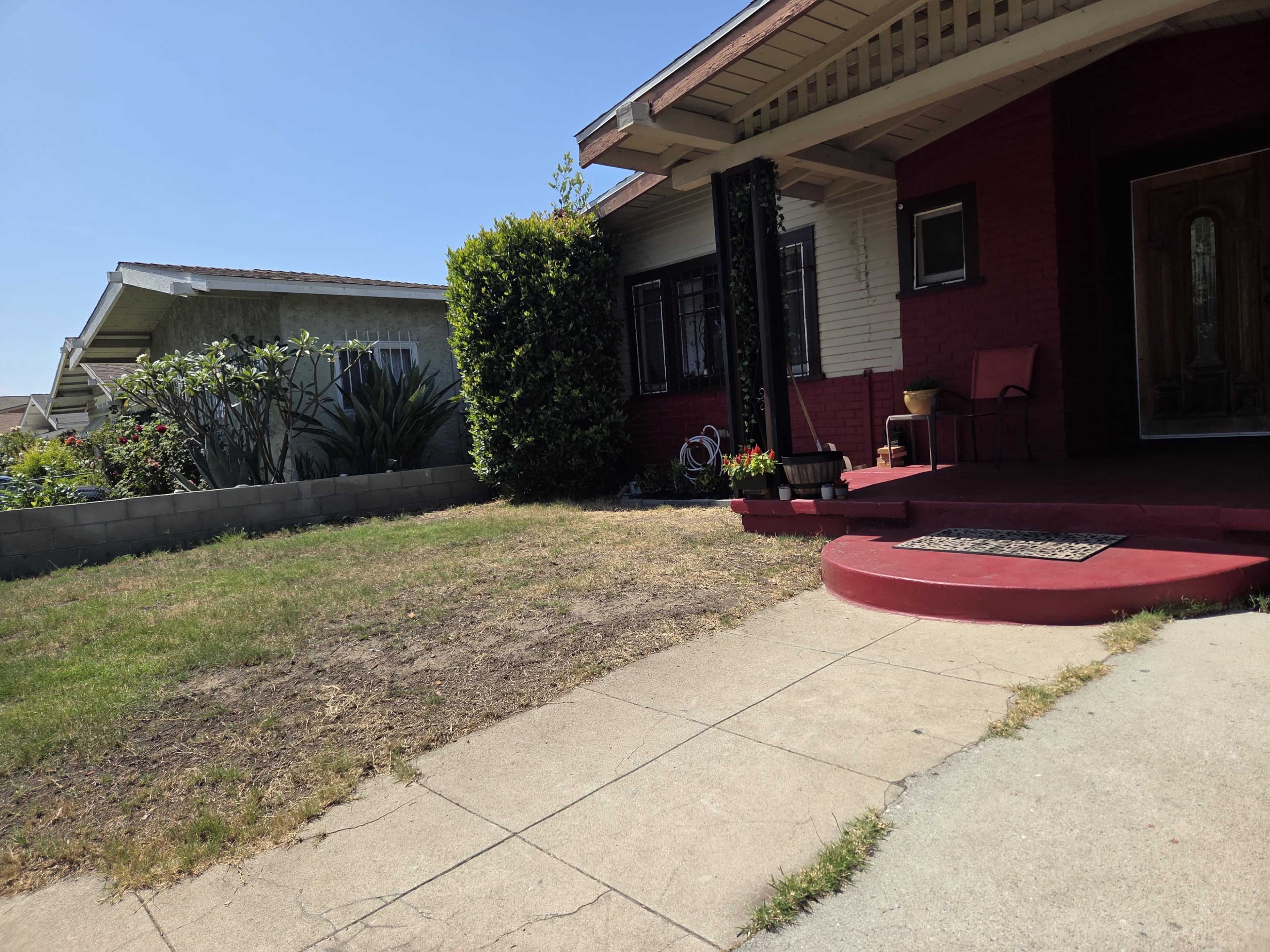 The image shows a front yard of a house with a concrete pathway leading to a porch, bordered by grass and shrubs.