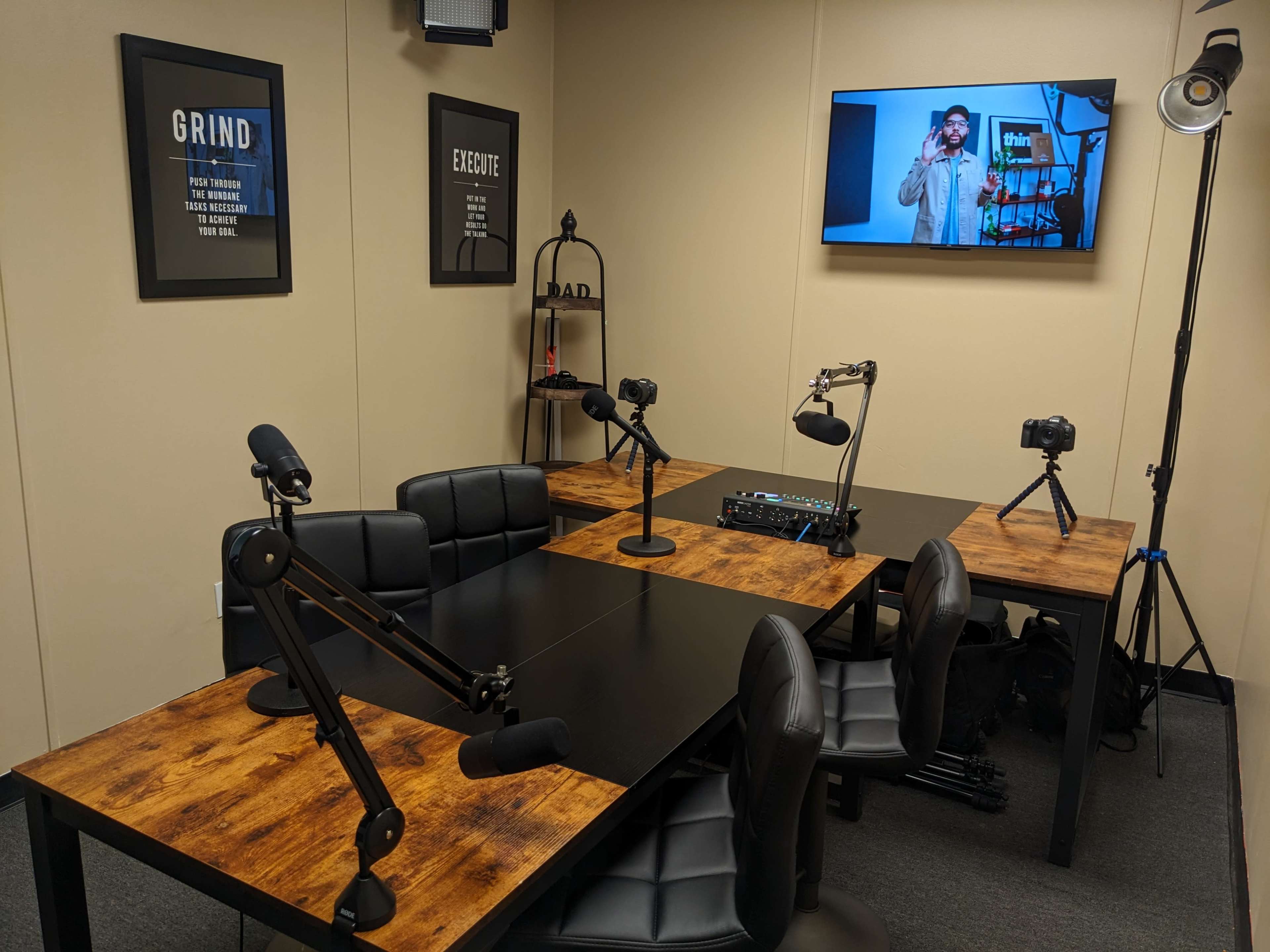 A podcast recording studio featuring two tables with microphones and cameras, a television screen displaying a person, and framed motivational posters on the walls.