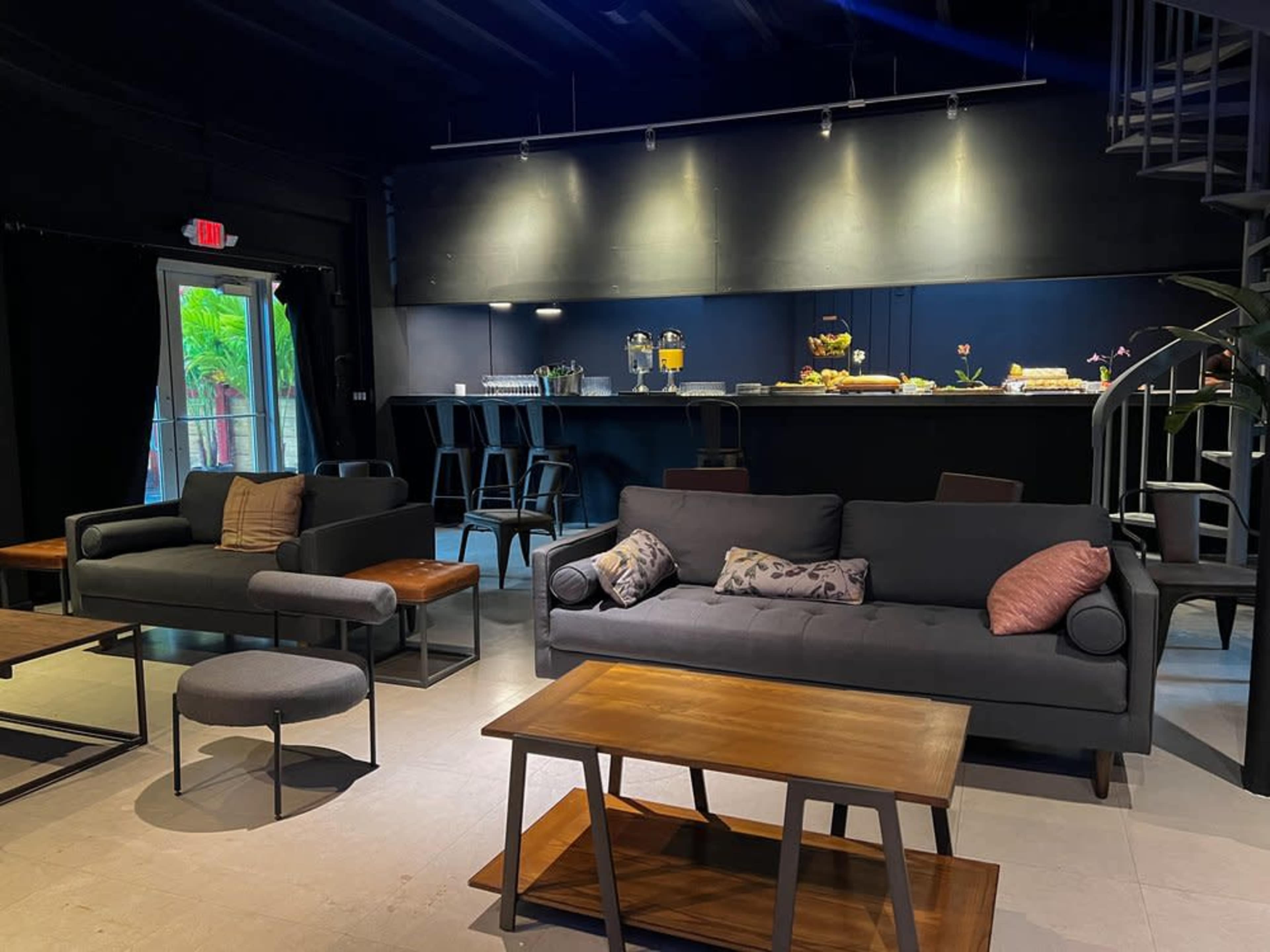The image shows a modern lounge area with gray sofas, wooden tables, and a bar counter in a dimly lit space.