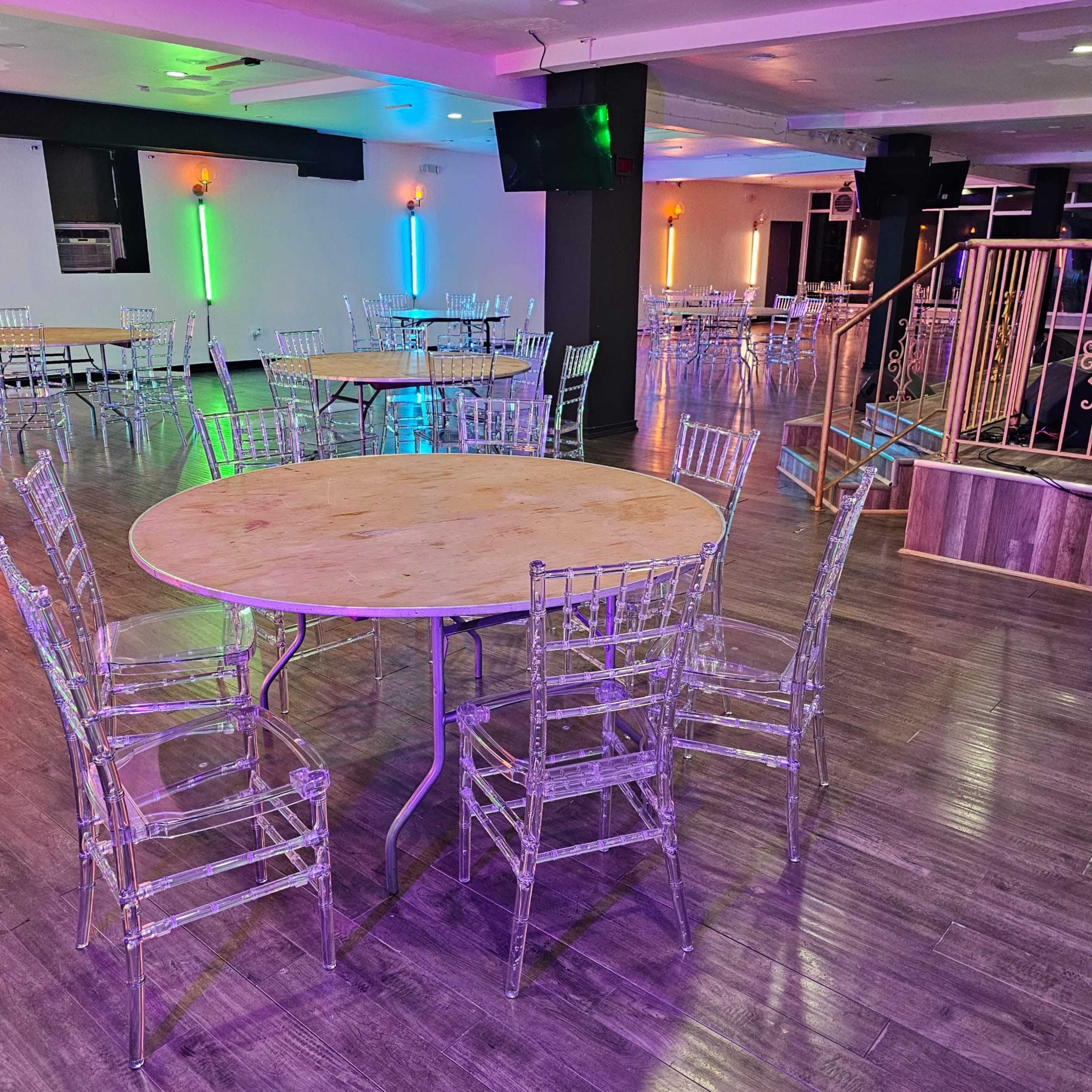 A large, empty event space features several round tables with transparent chairs arranged throughout, illuminated by colorful lights.