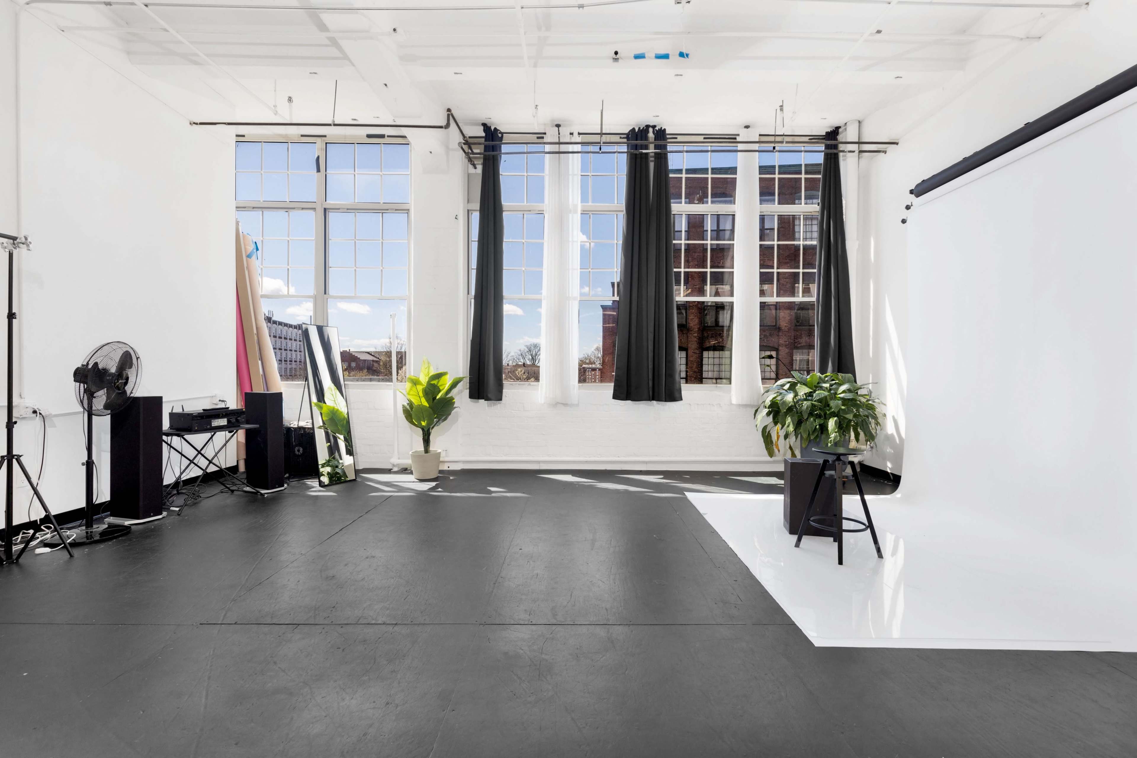 A bright, empty studio space with large windows, a black floor, and minimal furnishings, including a fan and potted plants.