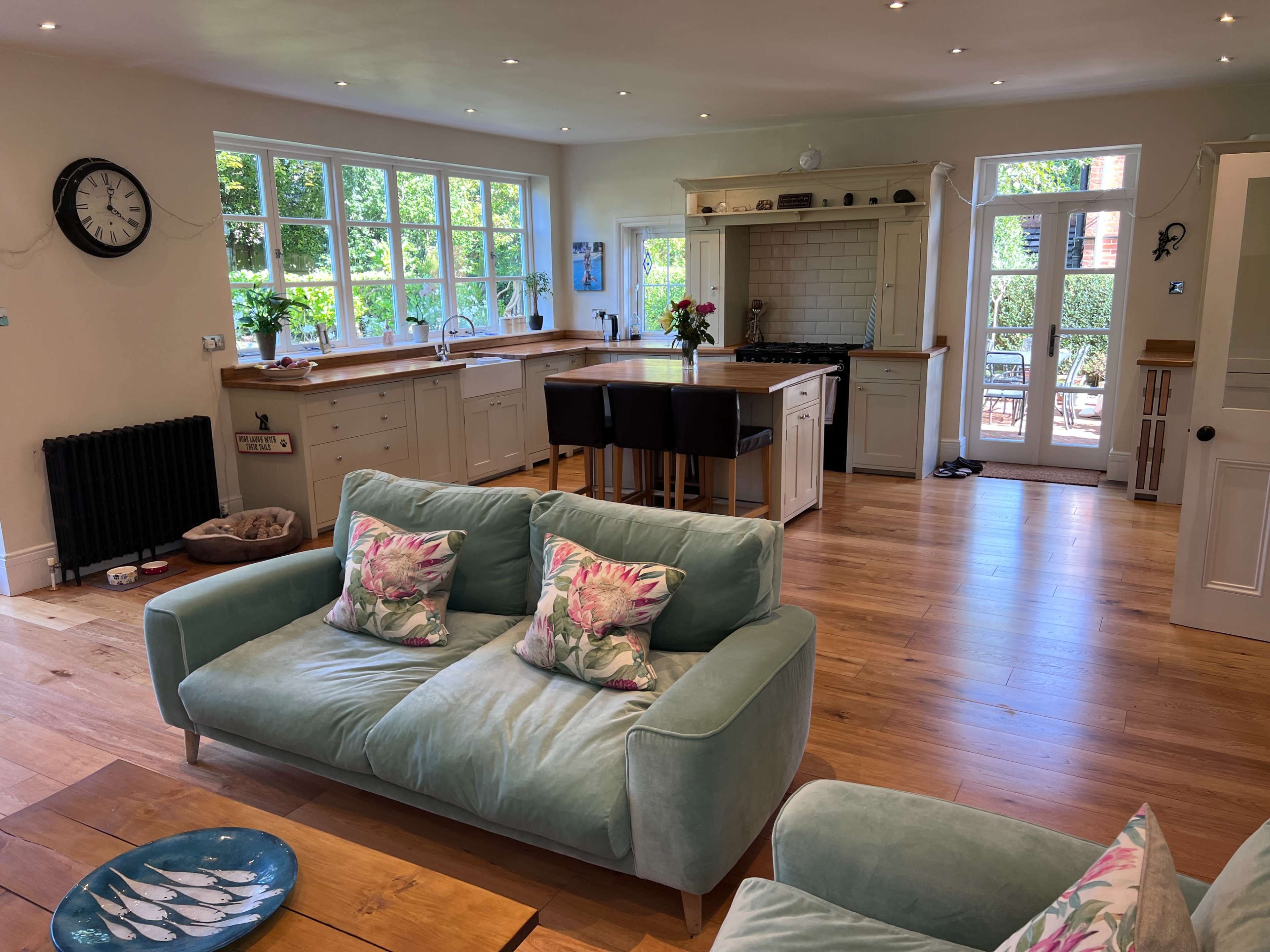 The image shows a bright, airy kitchen and living area with wooden floors, a large window, and a light-colored sofa with decorative cushions.