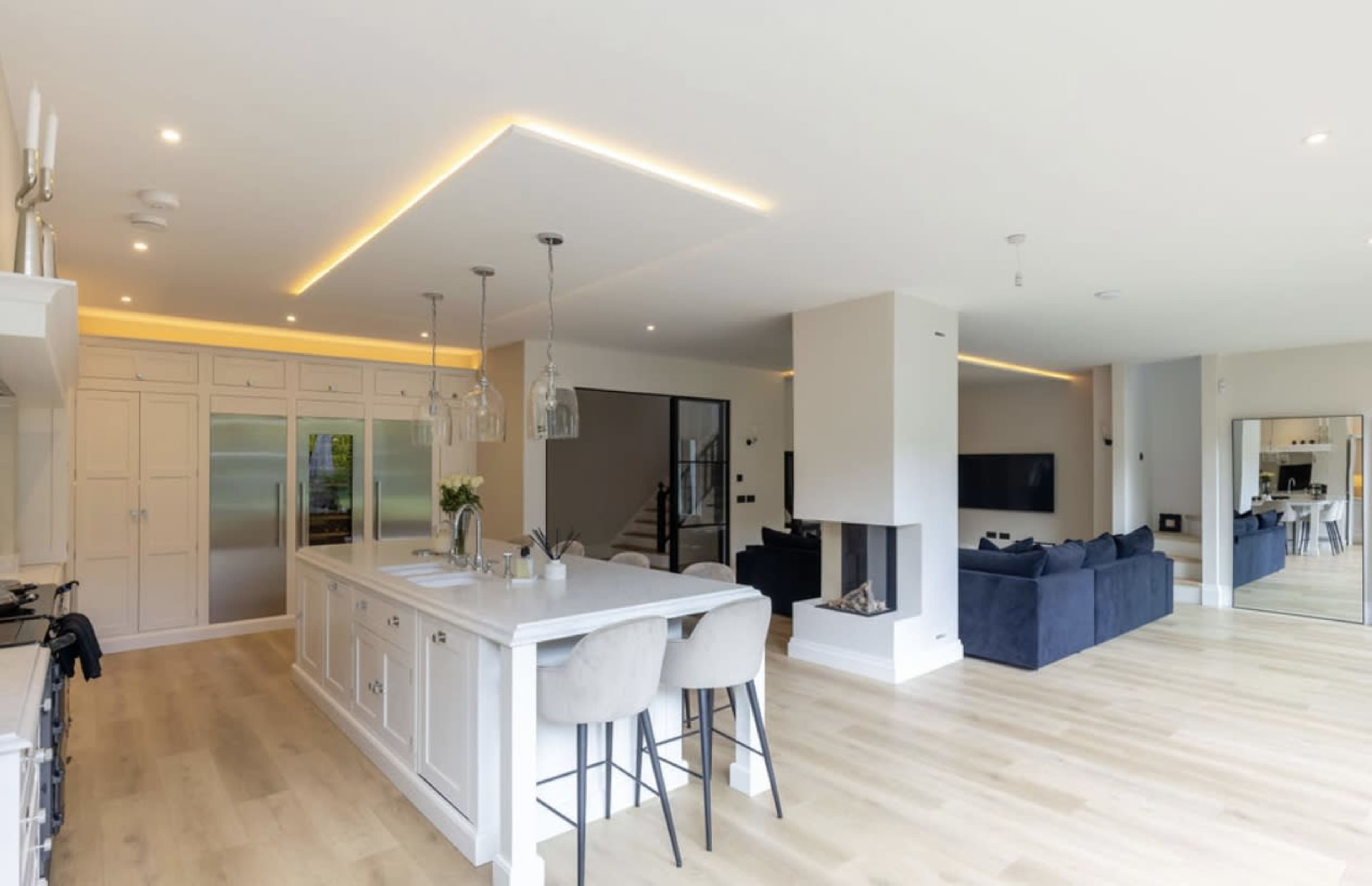 The image shows a modern open-concept kitchen and living area featuring a large island with bar stools, a fireplace, and a sleek design with ample natural lighting.