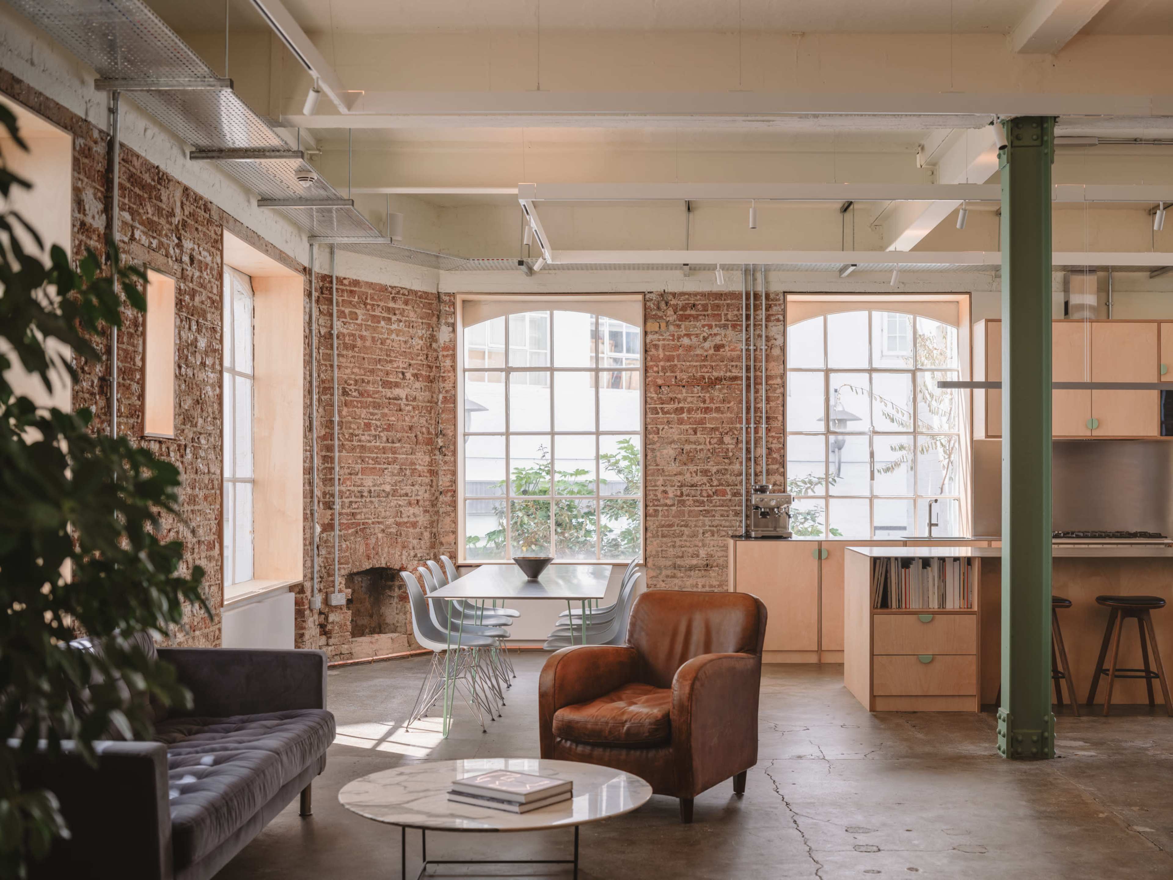 The image shows a modern, open-concept office space featuring exposed brick walls, a dining area with a table and chairs, and a lounge area with a leather armchair and a coffee table.