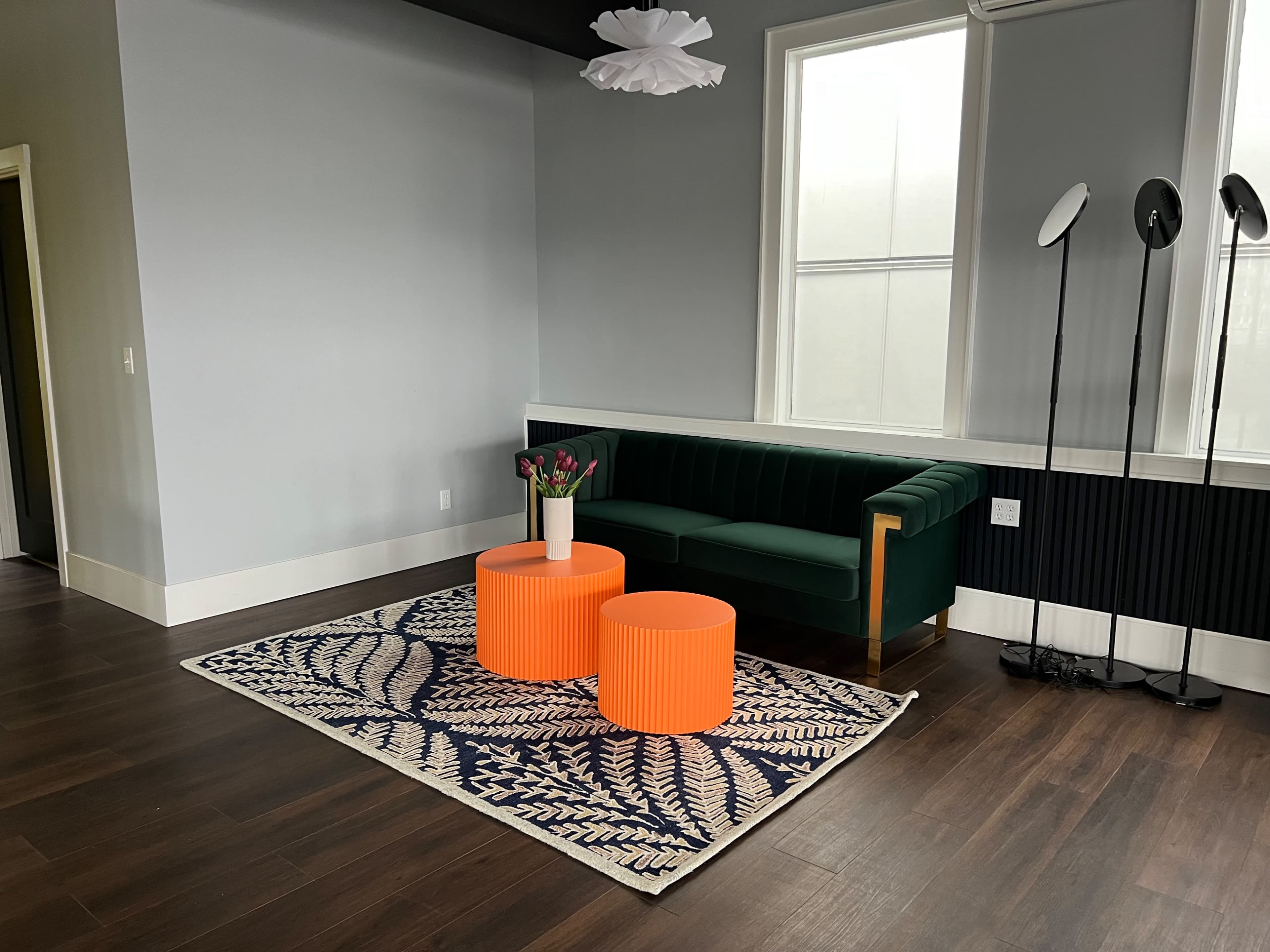The scene features a modern lounge area with a green sofa, two orange cylindrical tables, a flower arrangement, and a decorative rug on a dark wooden floor.