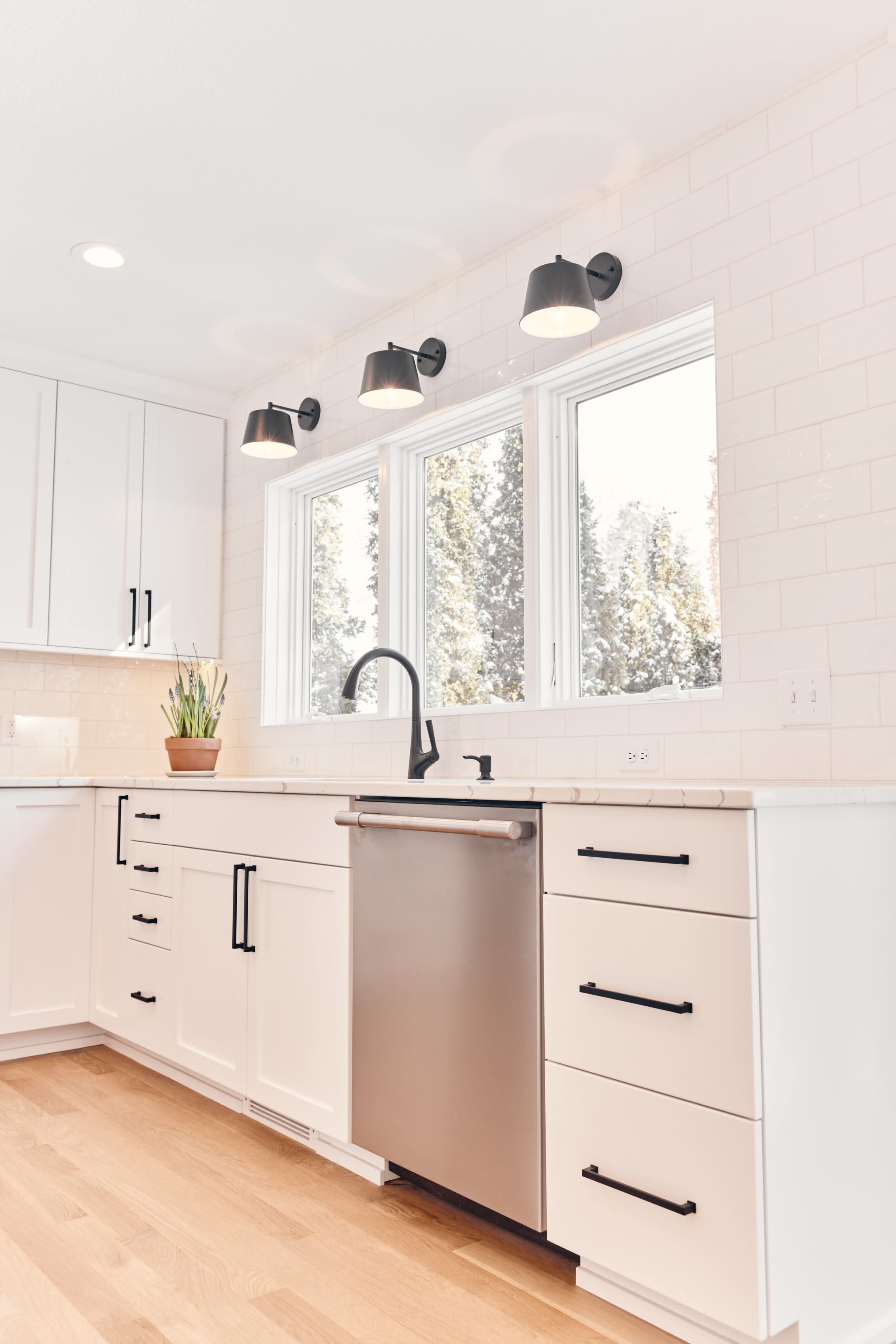 A modern kitchen features white cabinets, a stainless steel dishwasher, and three light fixtures above a sink with a window.