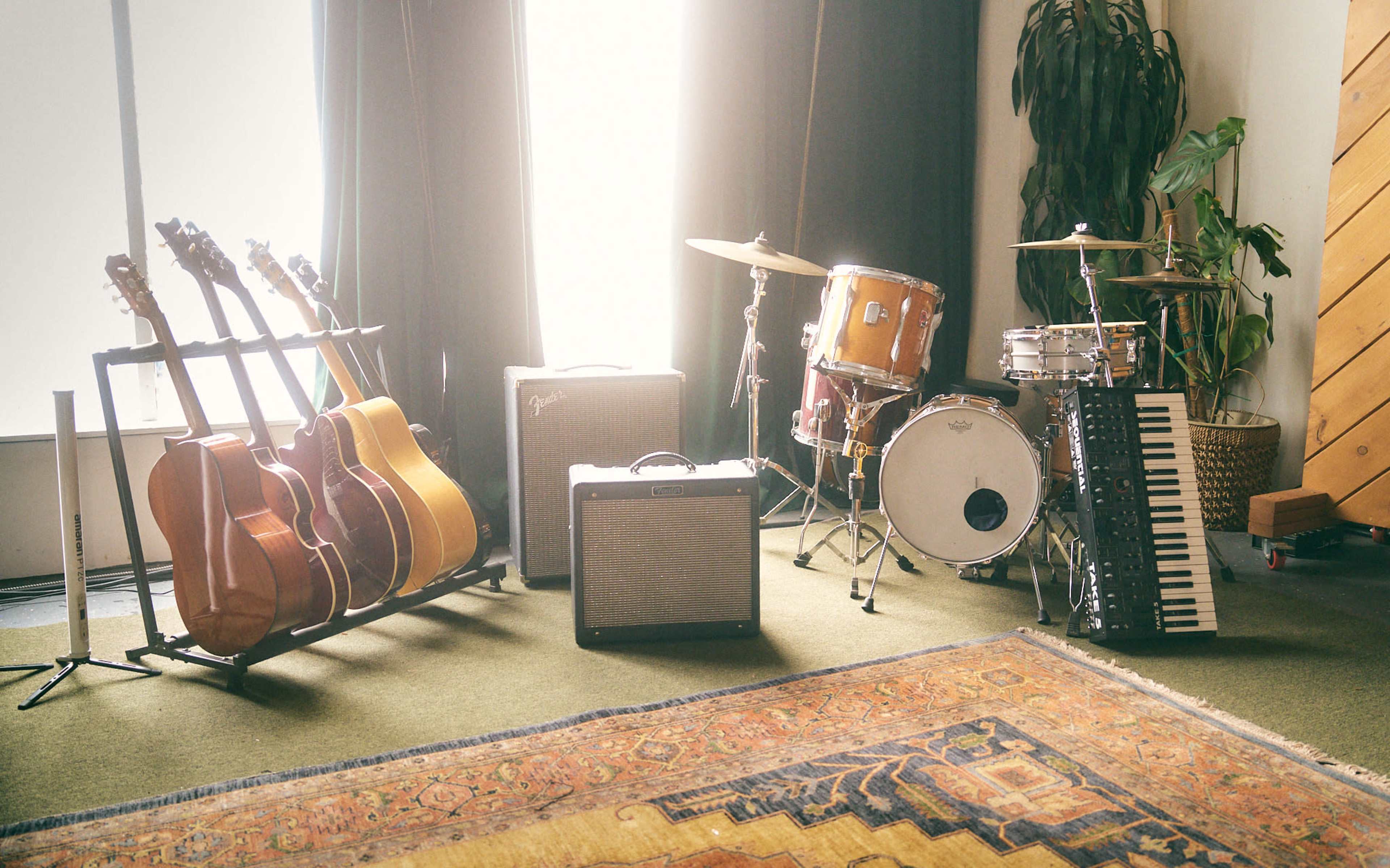 The image features a collection of musical instruments, including several guitars on a stand, a drum set, an amplifier, and a keyboard placed in a well-lit room with greenery and a patterned rug.