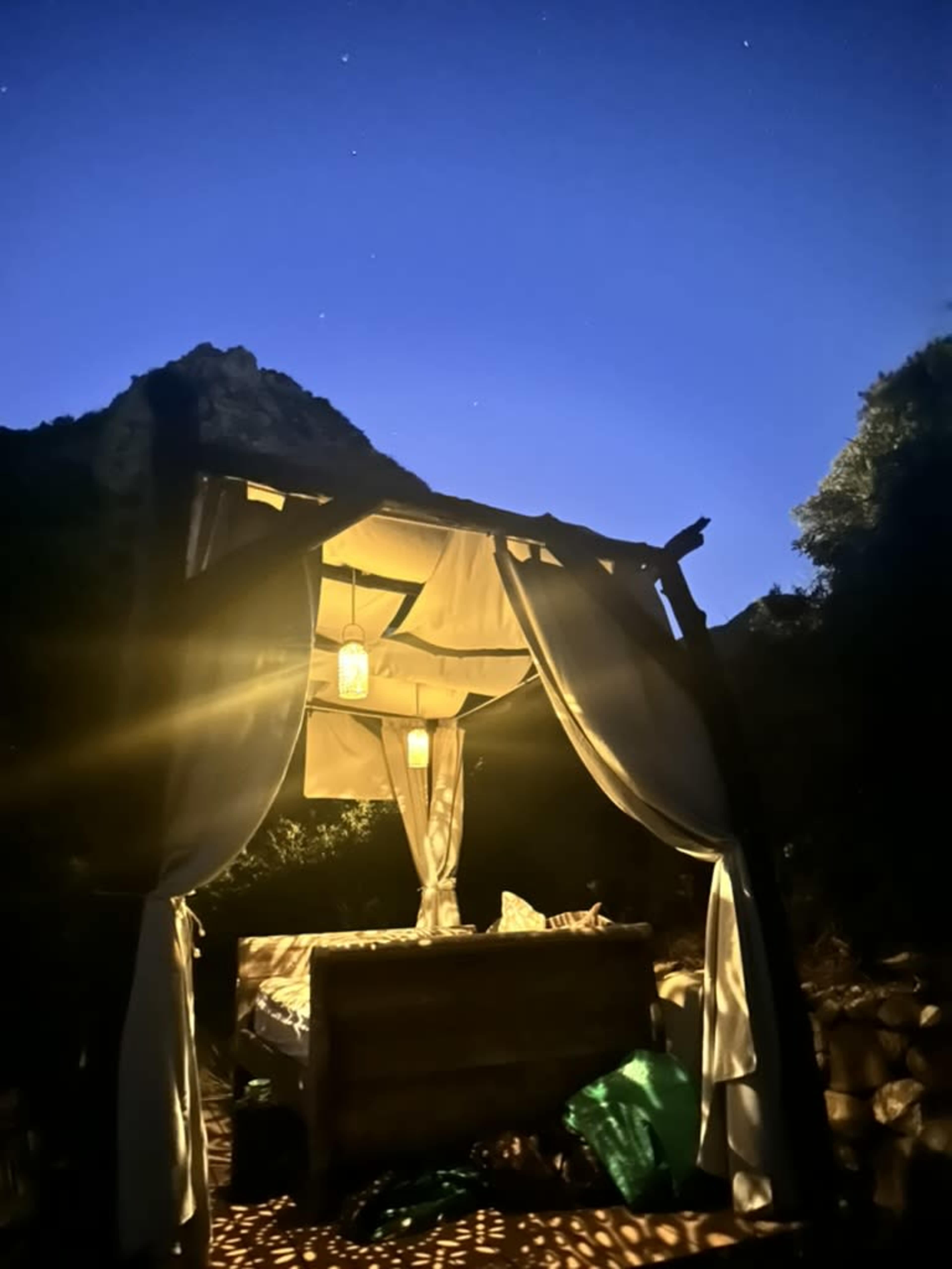 A wooden structure with draped fabric and lanterns sits in a dimly lit outdoor setting under a night sky.