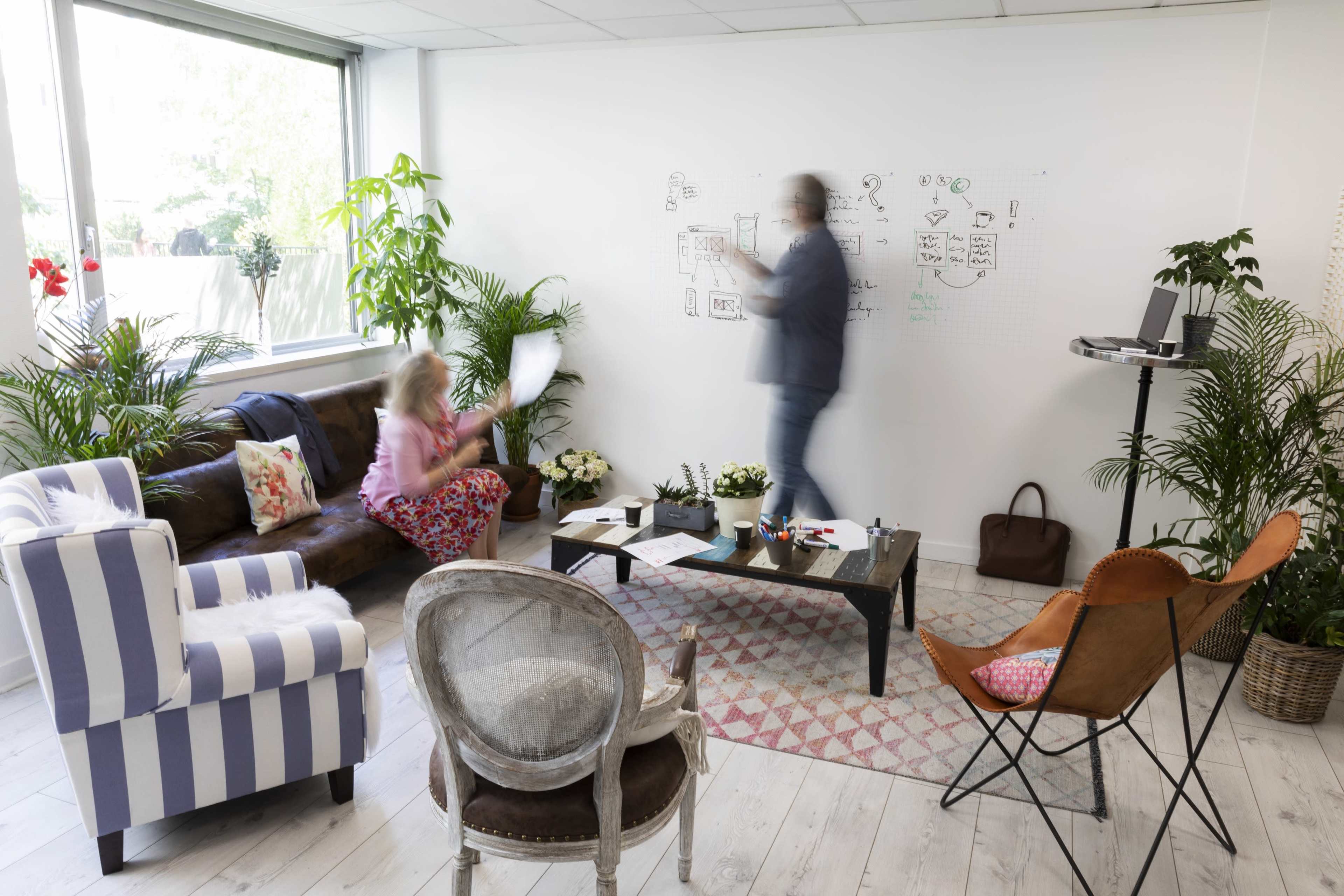 A person writes on a whiteboard while another sits on a couch holding a paper in a brightly lit, plant-filled office space.