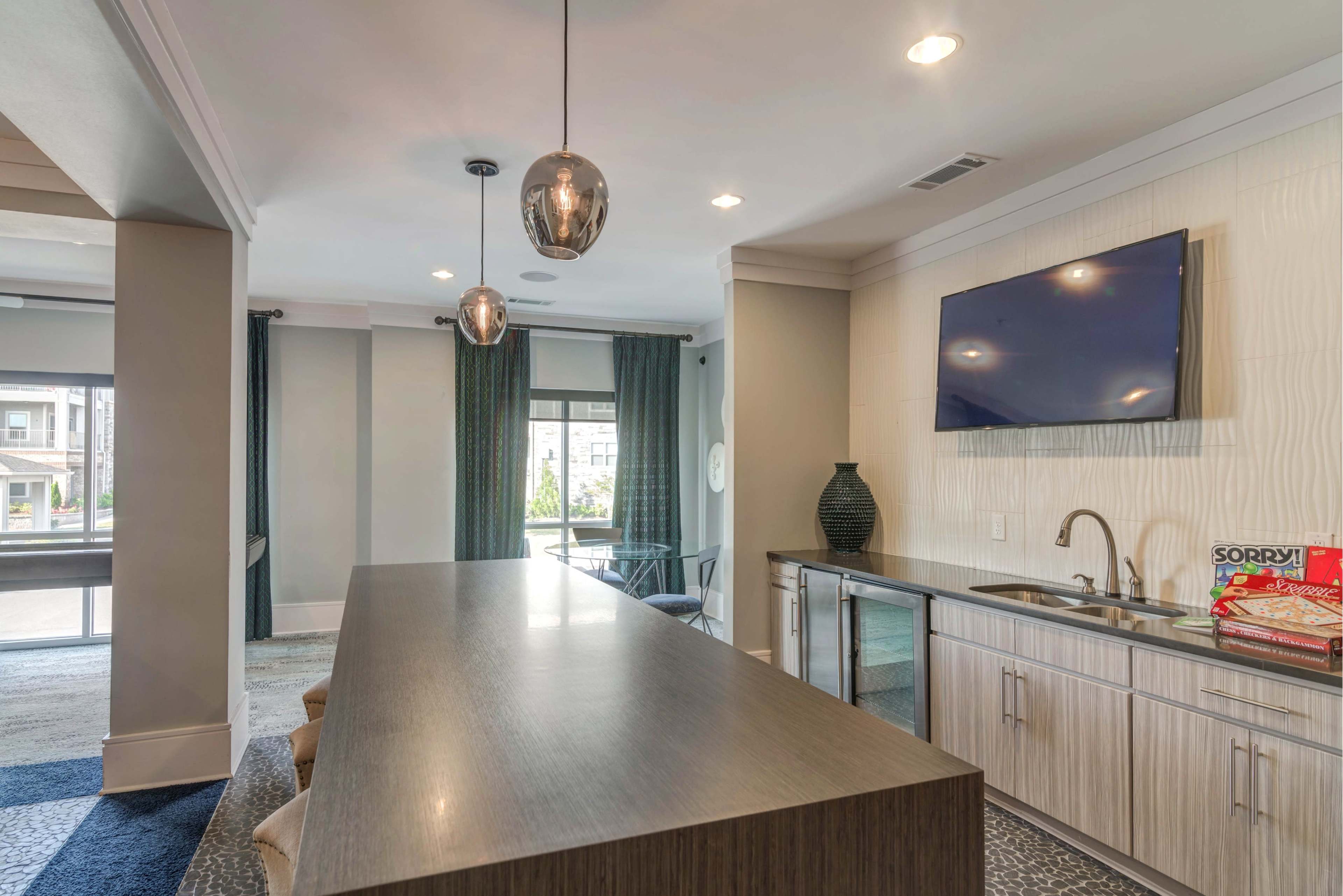 The image shows a modern lounge area featuring a long countertop with bar seating, a television mounted on the wall, and pendant lighting above.