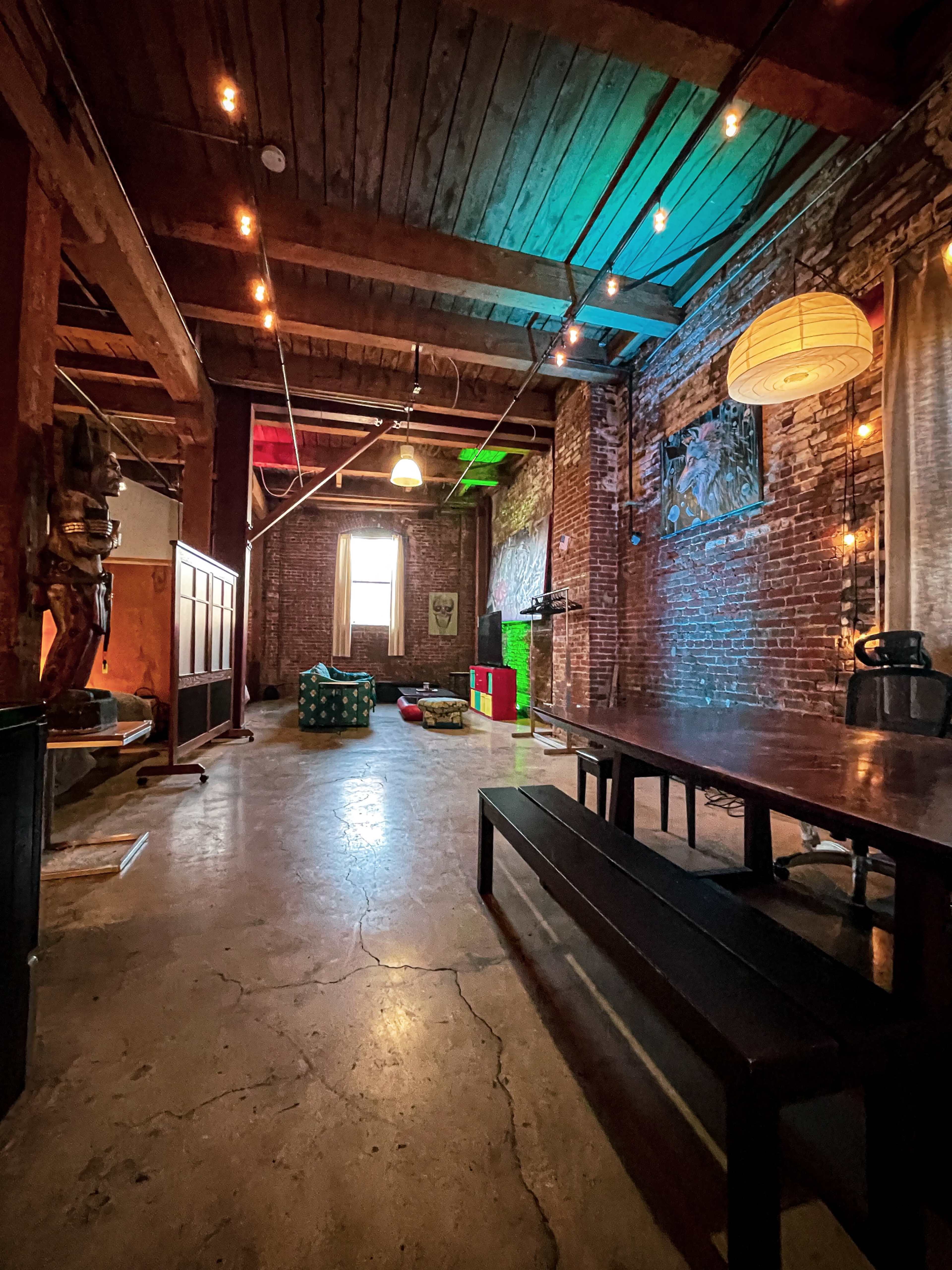 A spacious, industrial-style room with exposed brick walls, wooden beams, and various colorful decorations, including a large table and scattered seating areas.