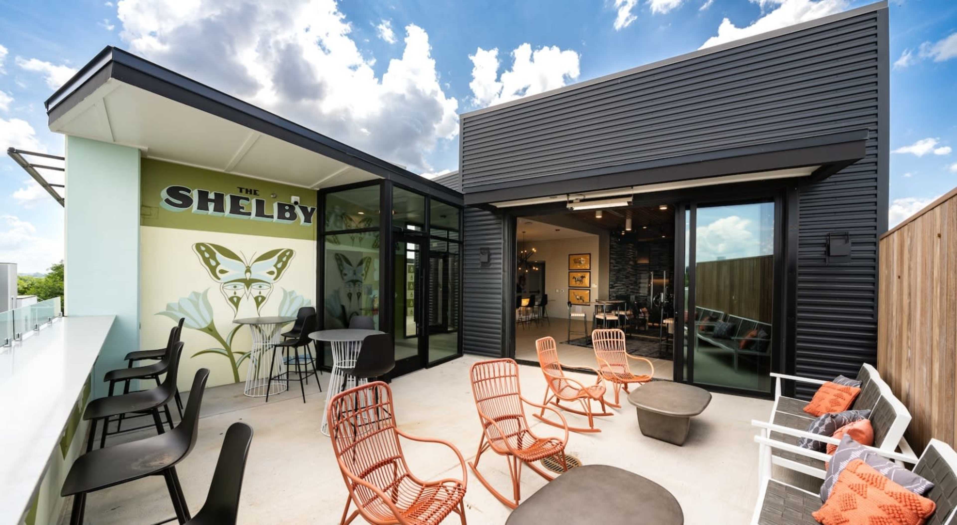 The image depicts a modern outdoor seating area featuring colorful chairs and tables, surrounded by large glass walls, with a mural and a cloudy sky in the background.