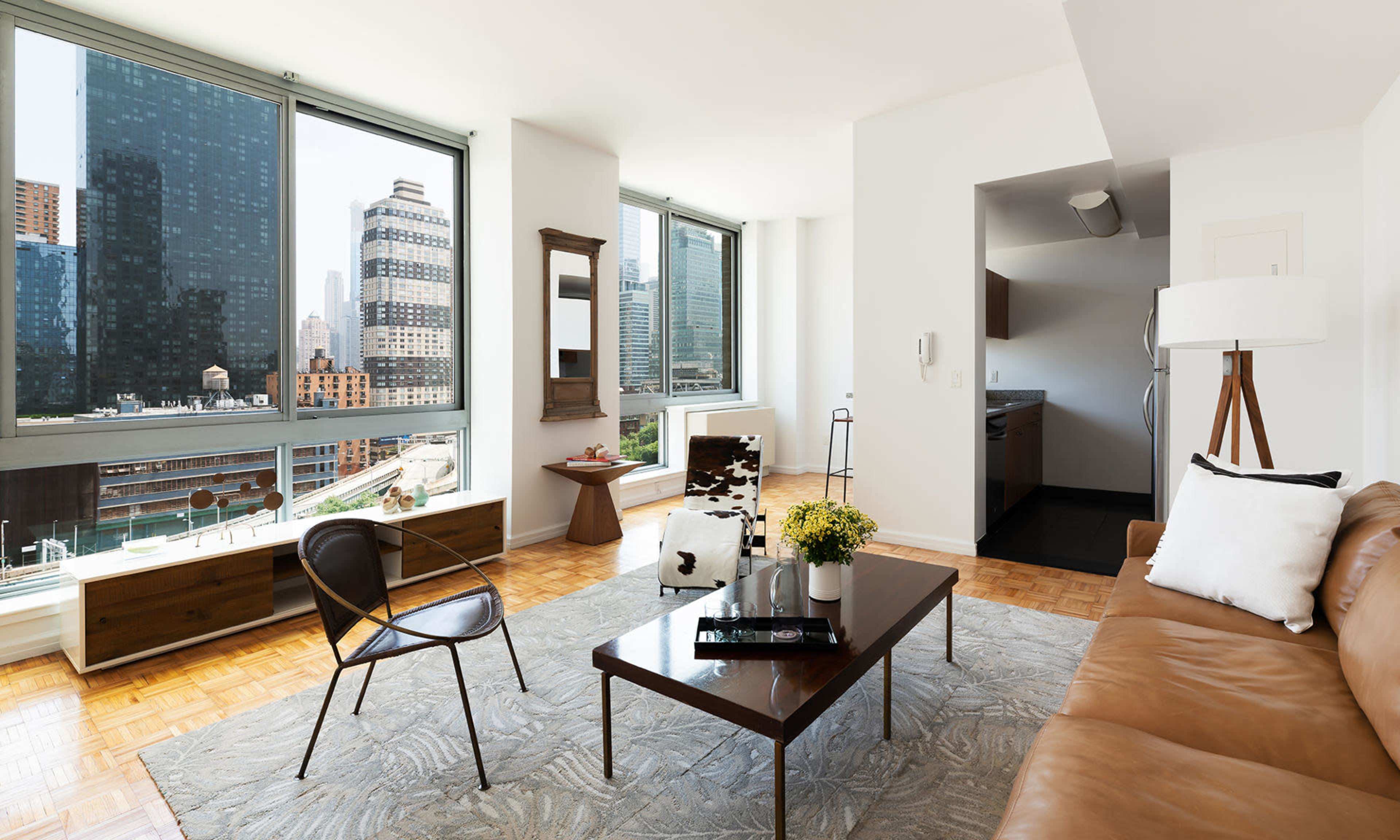 A modern living room features large windows with city views, a brown leather sofa, a coffee table, and decorative plants.