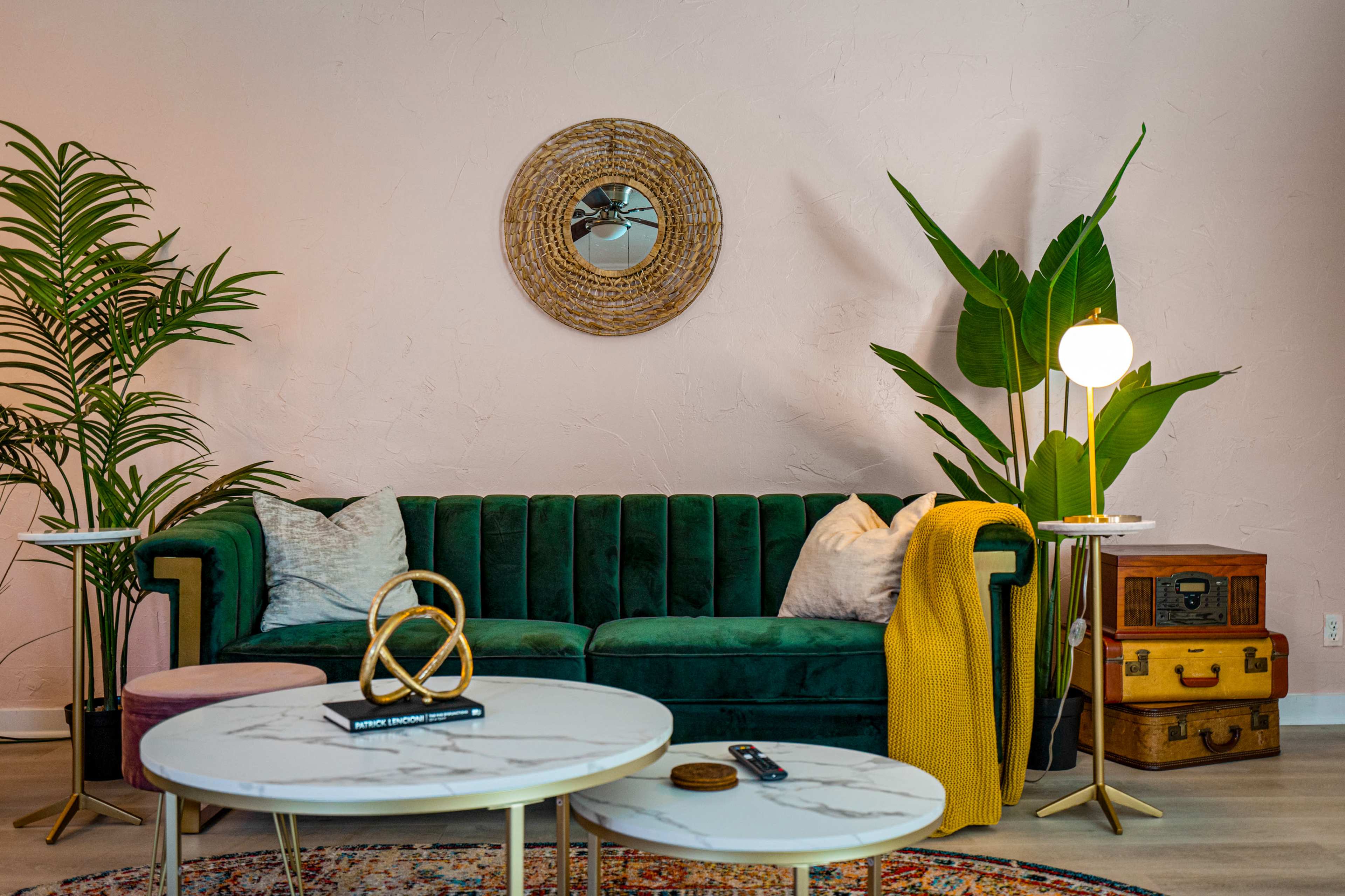 The scene shows a stylish living room featuring a green velvet sofa, a round mirror, potted plants, a marble coffee table, and accent decor on a light pink wall.
