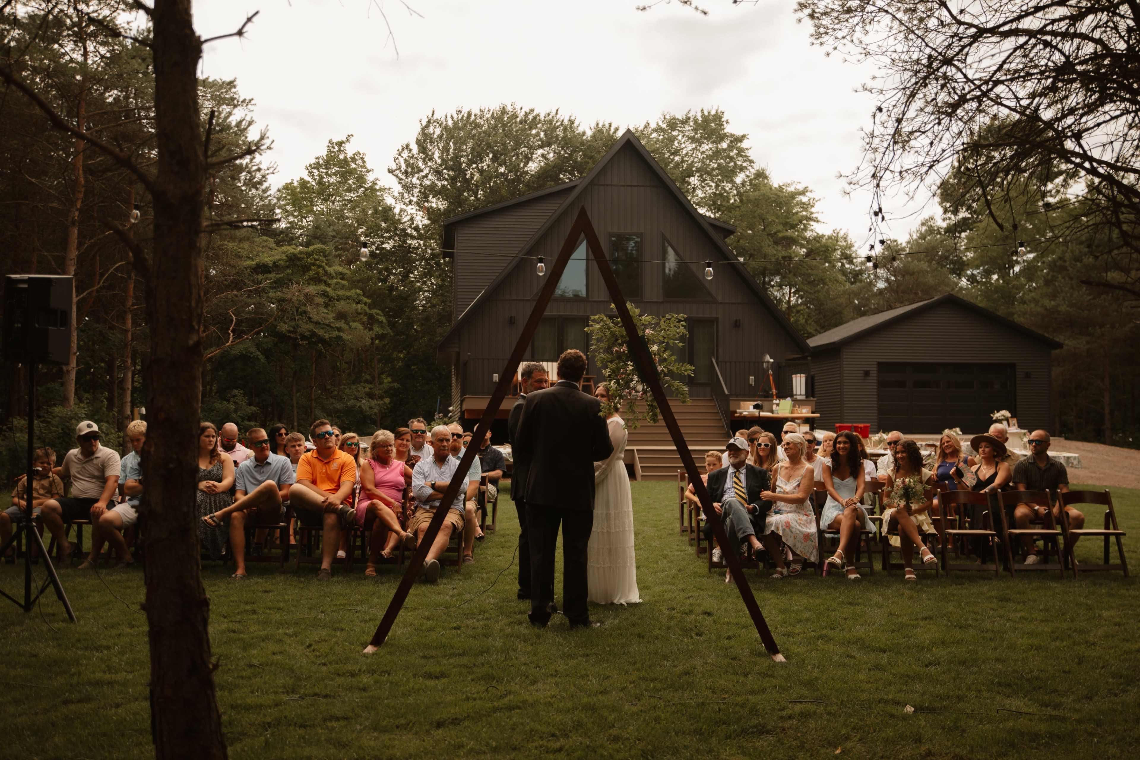 A couple stands under an arch while exchanging vows in front of a seated audience at an outdoor wedding ceremony near a dark-colored house.