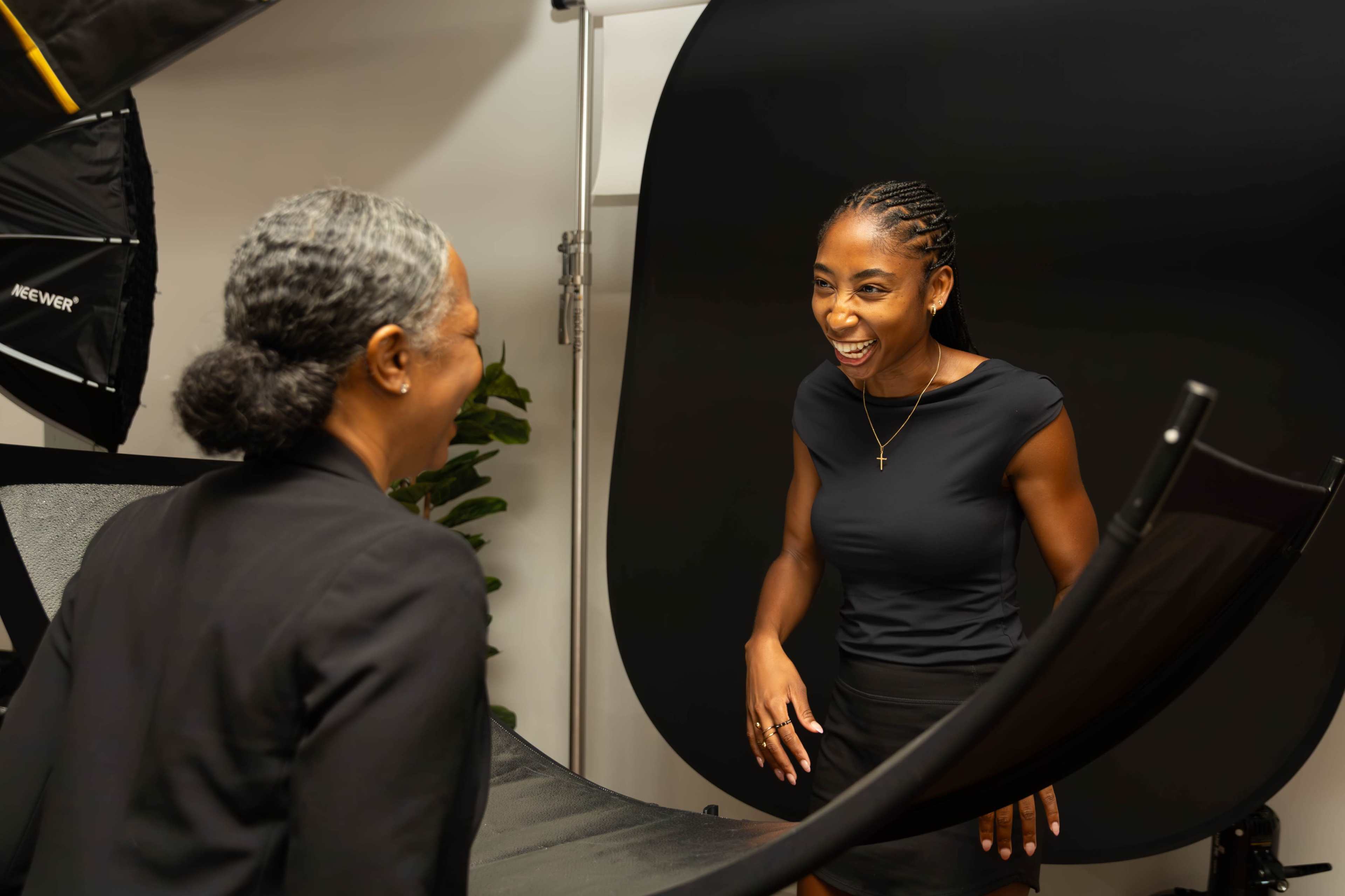 Two women are laughing and interacting in a photo studio with a dark backdrop and soft lighting equipment.