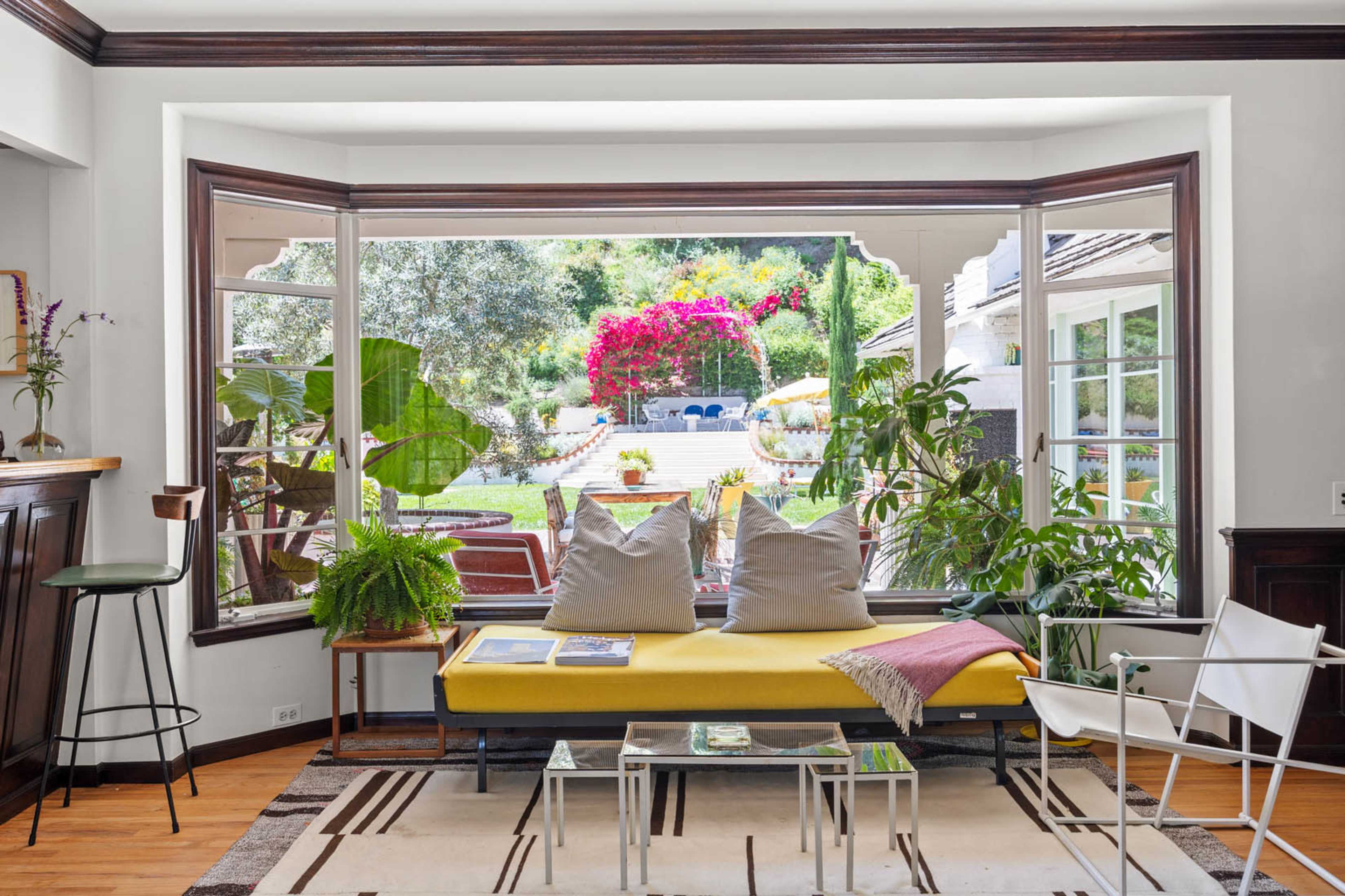 The image shows a cozy living room with a yellow sofa and large windows that open up to a landscaped garden featuring colorful flowers and outdoor seating.