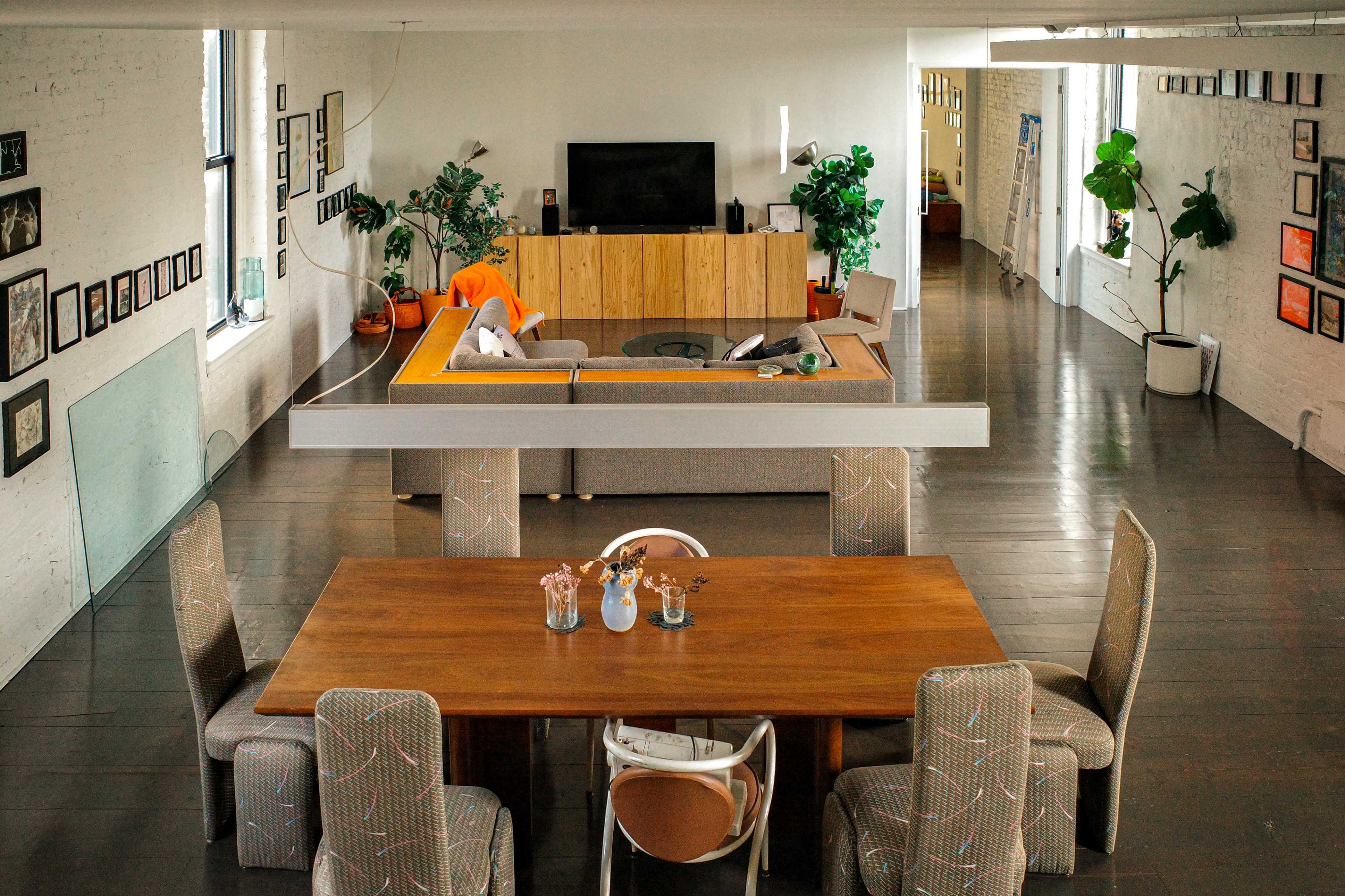The image shows a spacious and modern interior with a large dining table, surrounding chairs, a living area with a sectional sofa, and a television on a wooden console.
