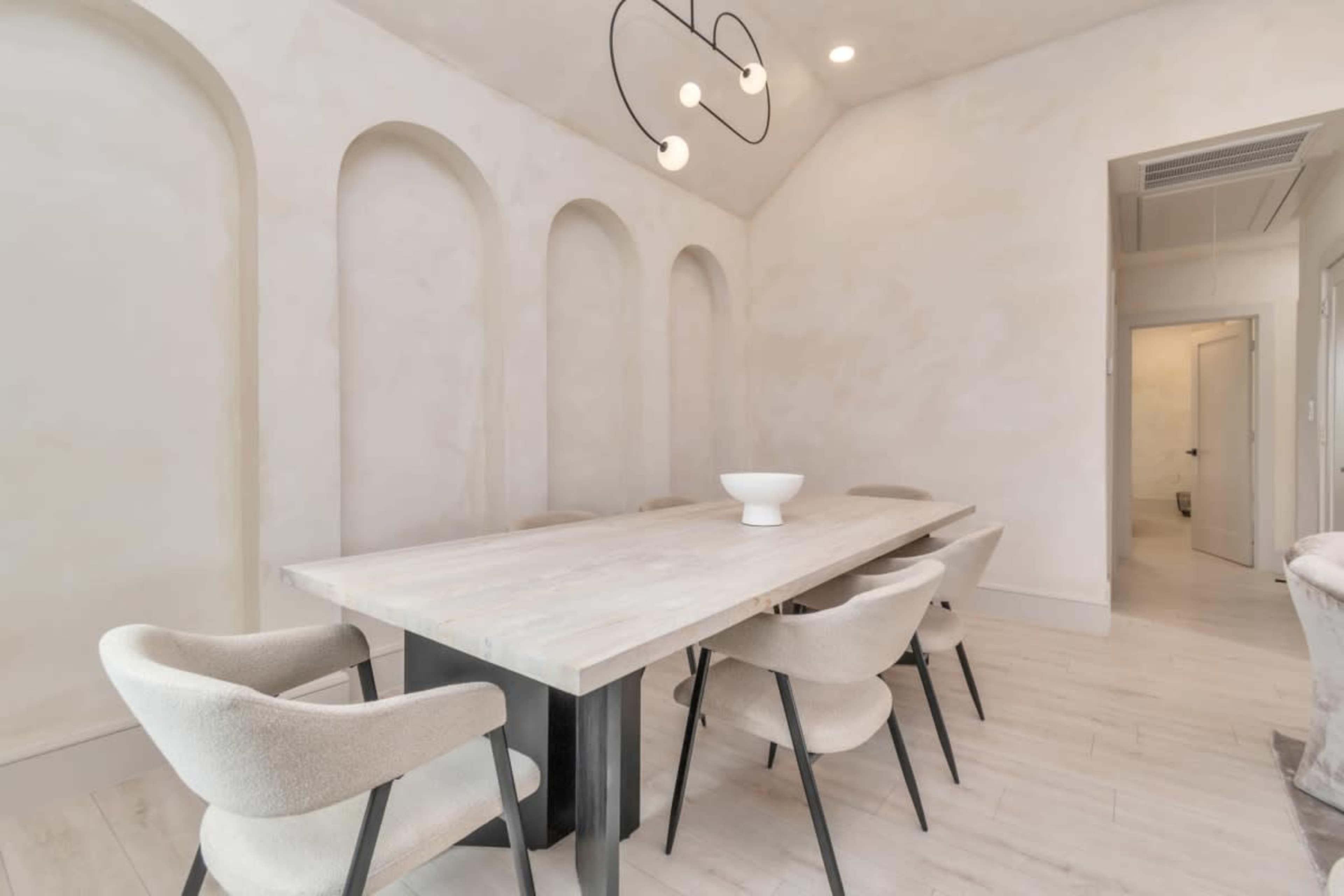 A minimalist dining room features a long wooden table surrounded by upholstered chairs beneath a modern light fixture and accented by curved wall niches.