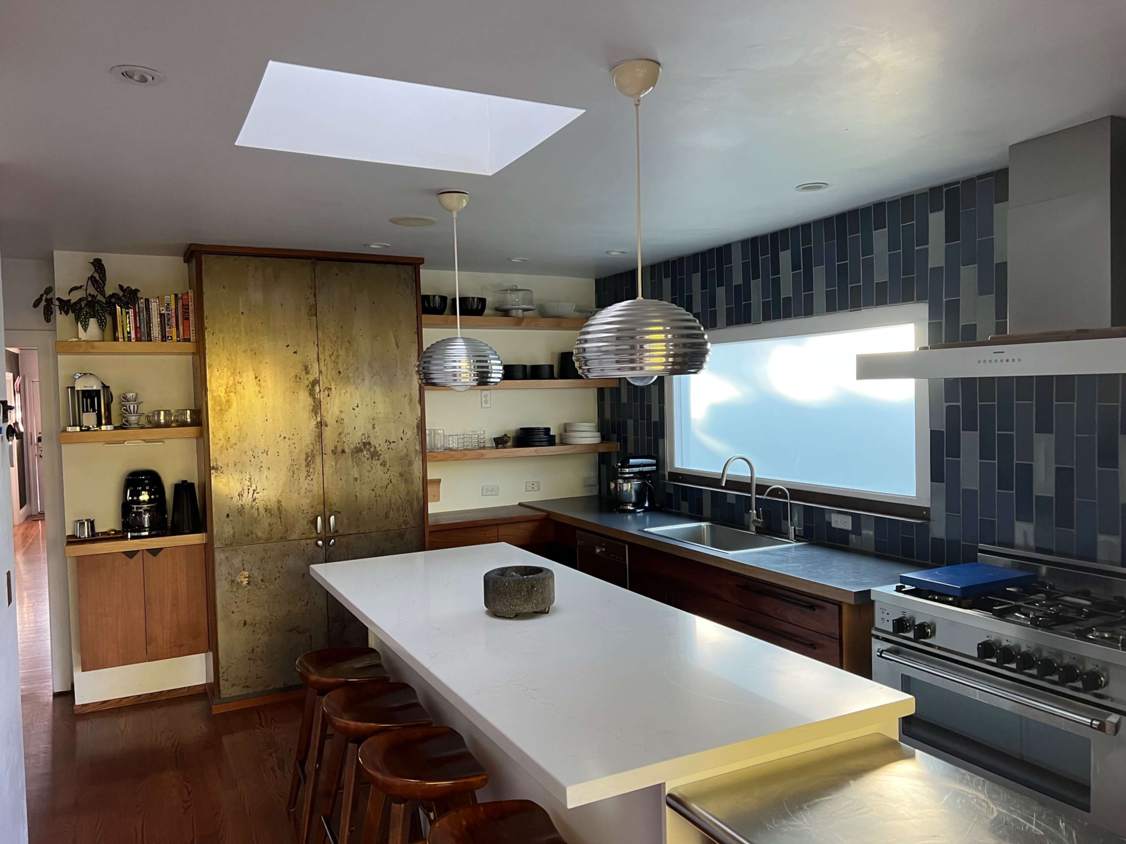 The kitchen features a large island with four stools, sleek appliances, and a mix of wooden and tiled finishes, all illuminated by natural light through a skylight.