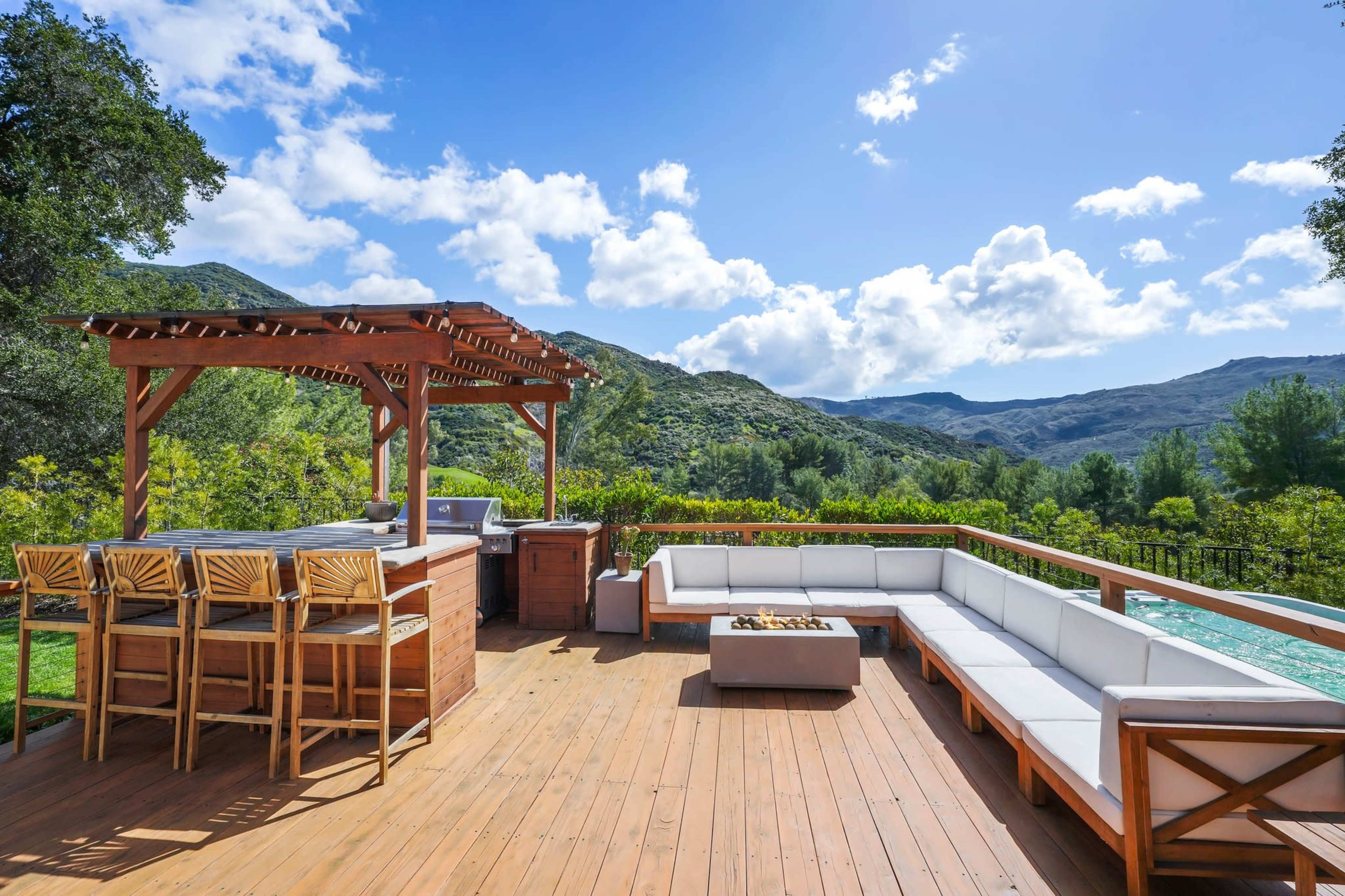 The image features a spacious wooden deck with a lounge area and an outdoor bar overlooking a green landscape and mountains under a partly cloudy sky.