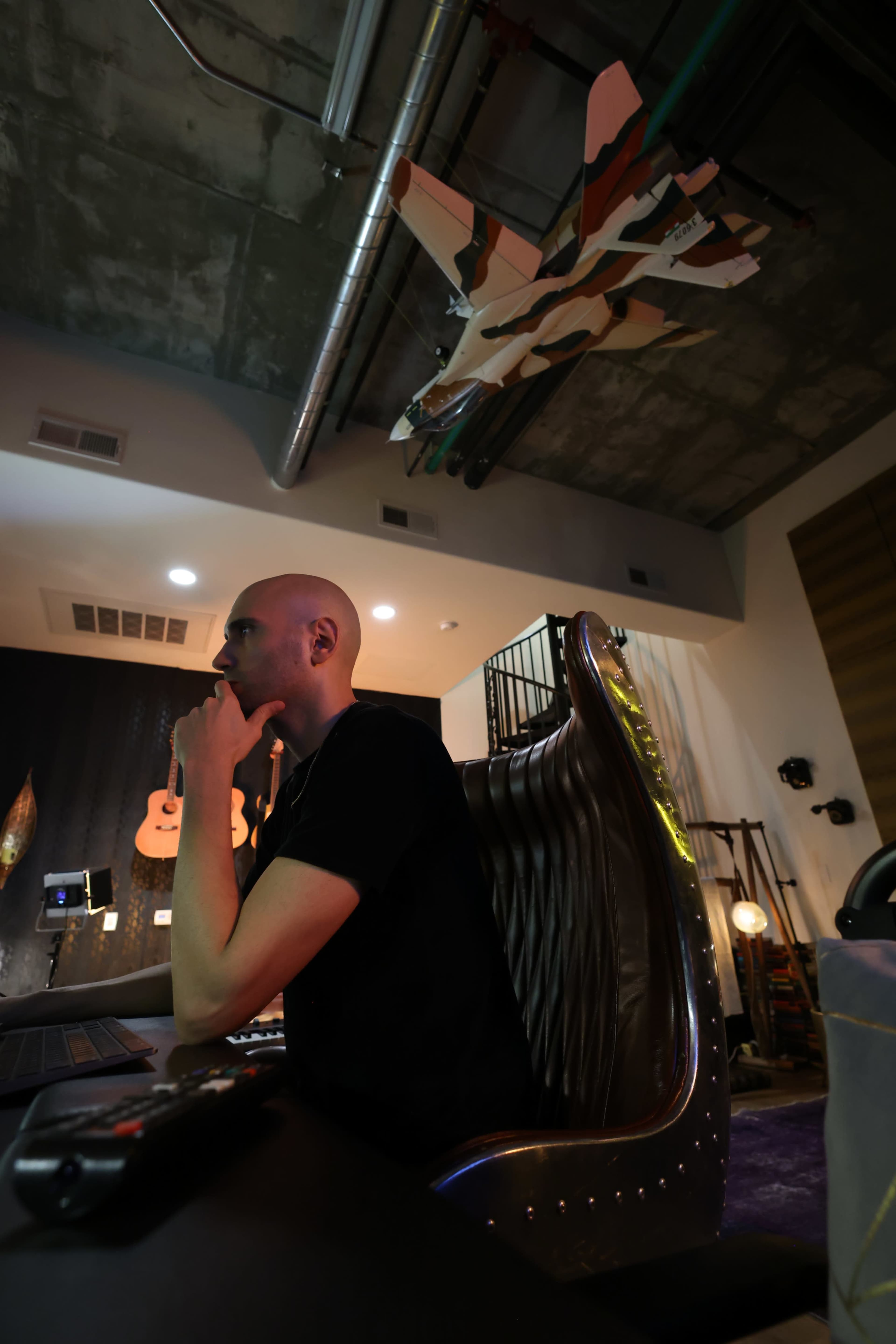 A person sits at a computer in a modern workspace, surrounded by a minimalist decor featuring guitar silhouettes and a ceiling installation of model aircraft.