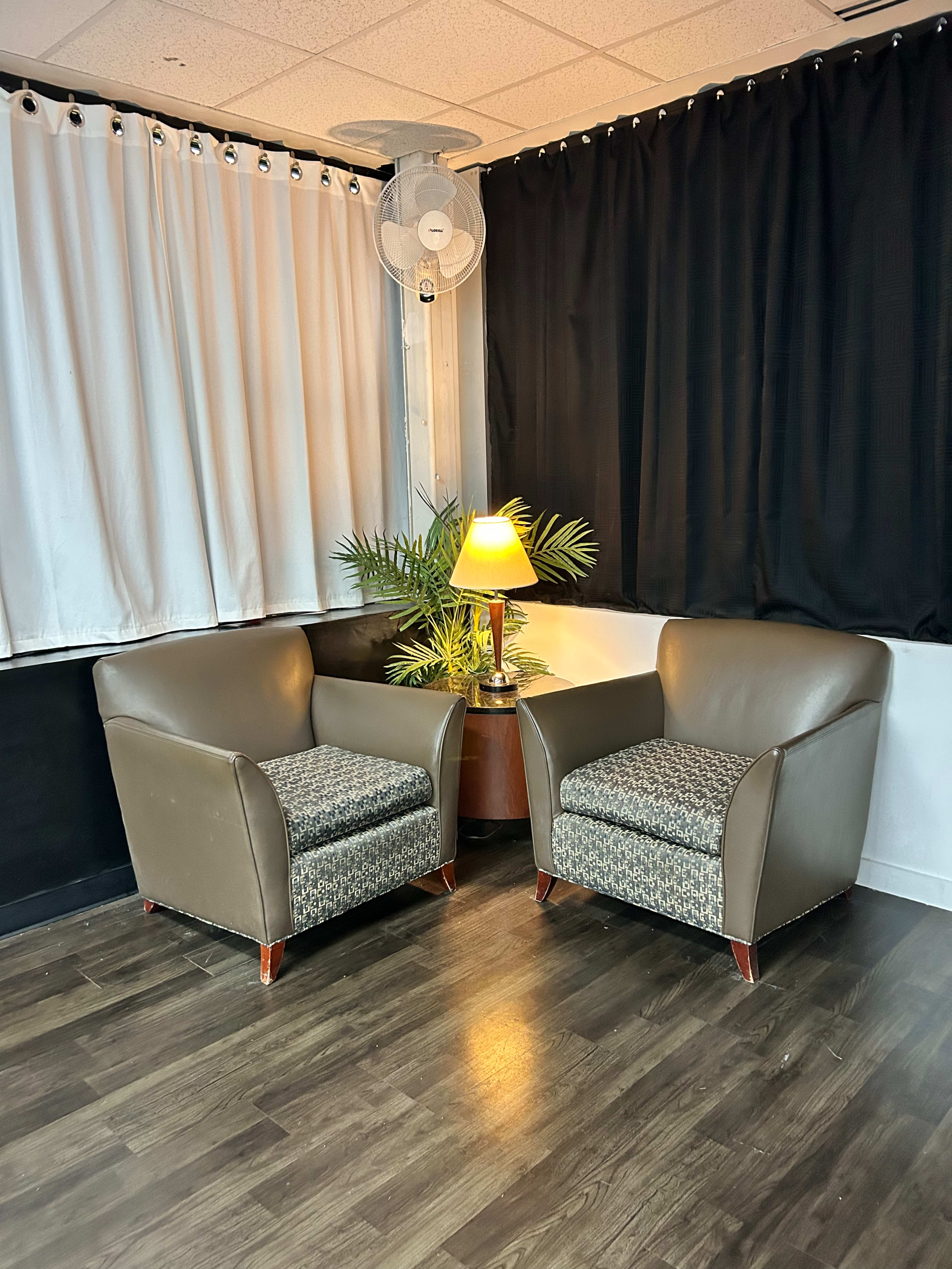 Two upholstered armchairs flank a small wooden table with a lamp and a potted plant in a room with black and white curtains.