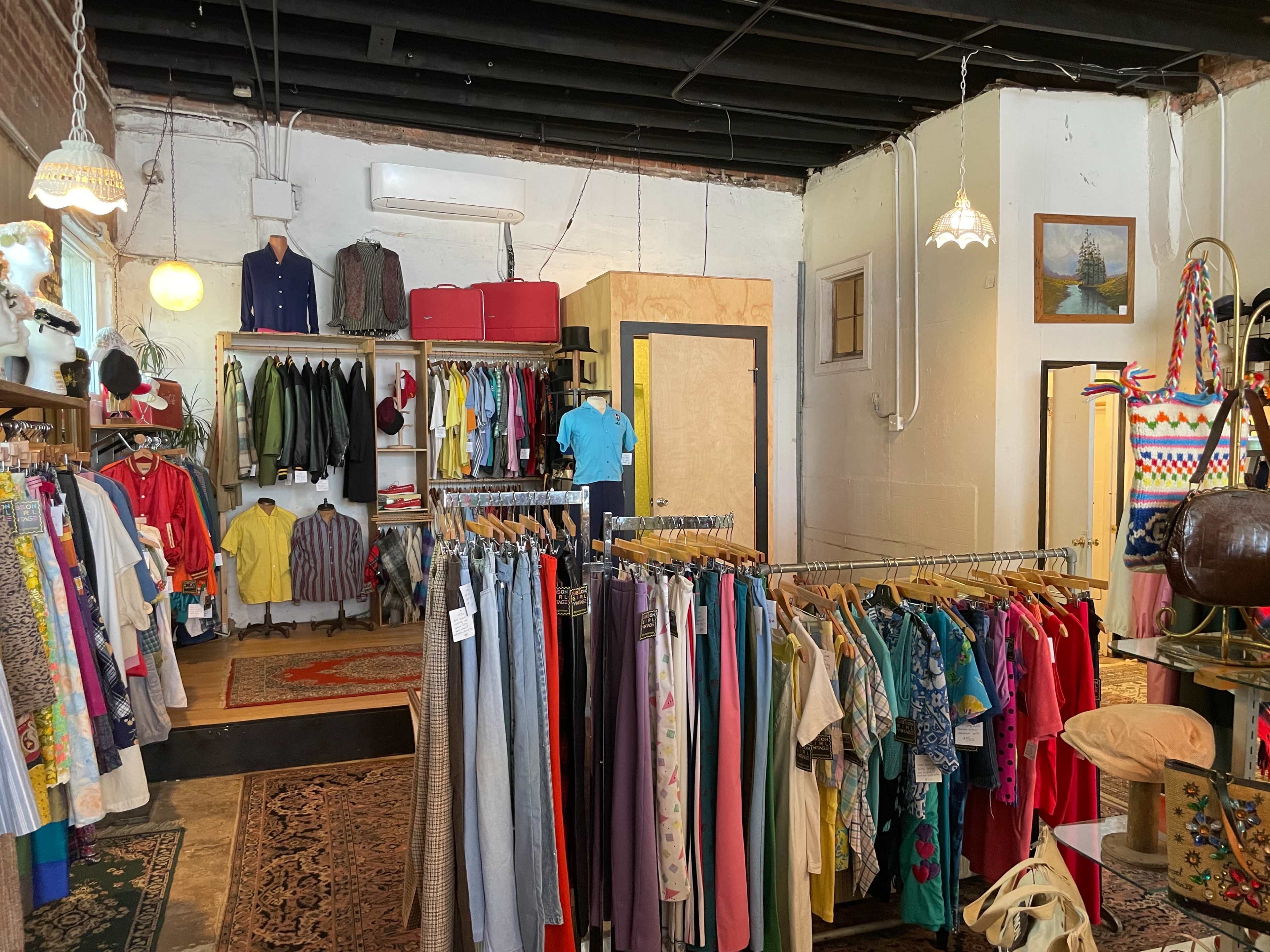 The interior of a vintage clothing store features racks of colorful garments, a cozy rug, and displays of various clothing items and accessories.